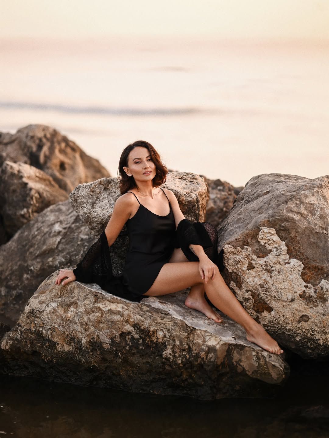 Female beach photoshoot in Valencia, with a woman in a black slip dress sitting gracefully on large coastal rocks at sunset.