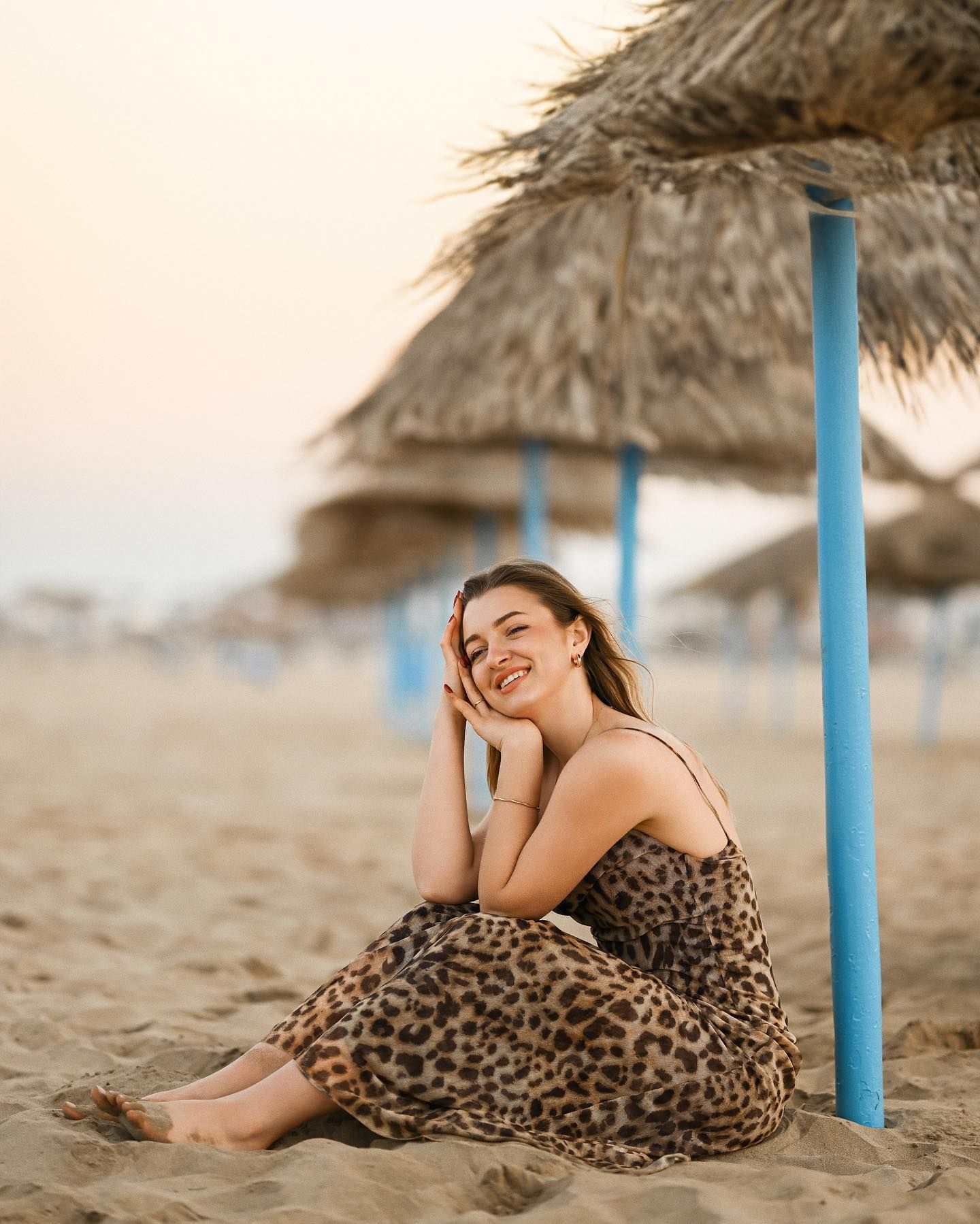 A smiling woman sitting on the sand next to straw beach umbrellas during a golden hour photoshoot in Alicante.