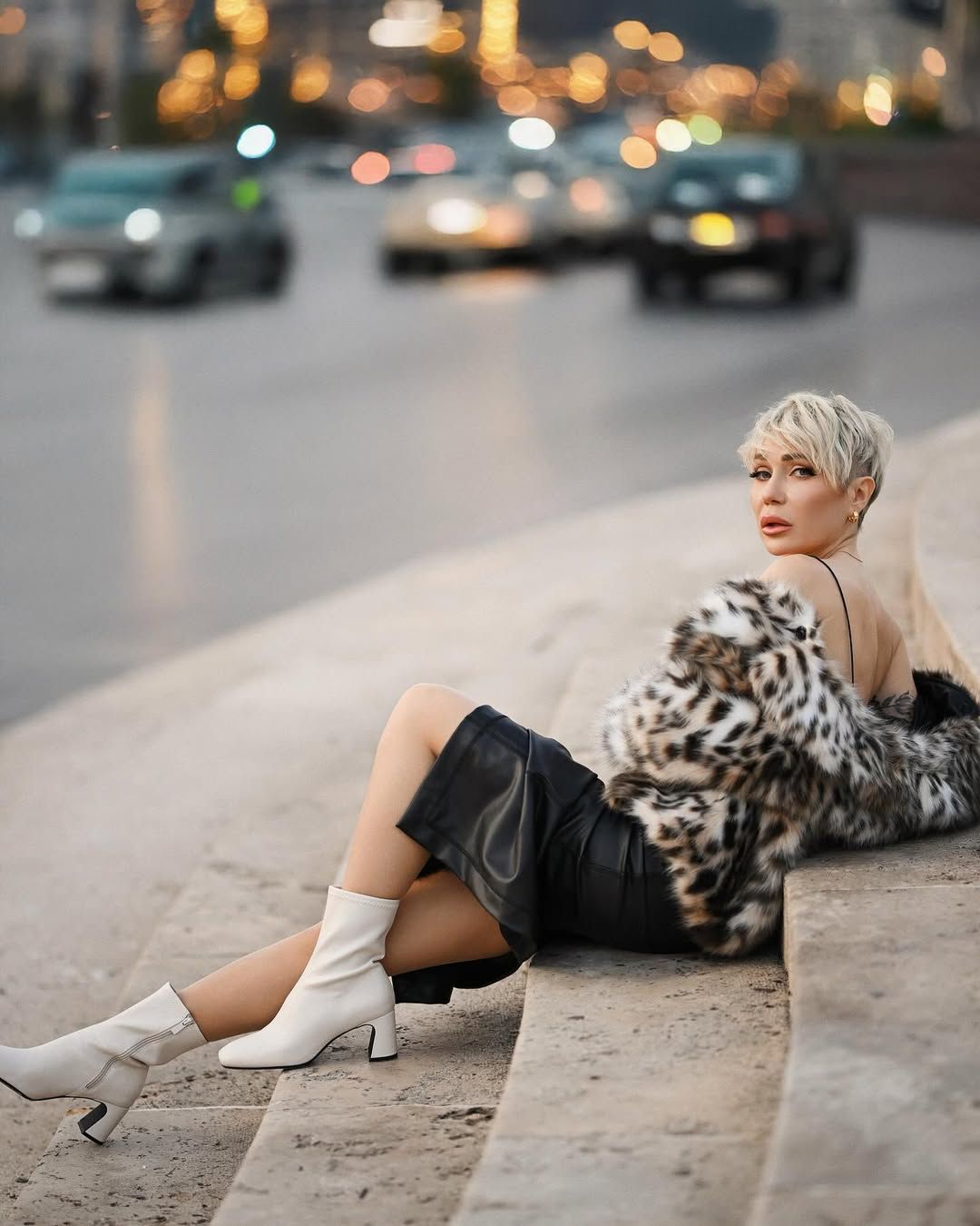 A luxury blonde woman in a curated outfit and white boots posing on stone steps during an all-inclusive outdoor photoshoot.