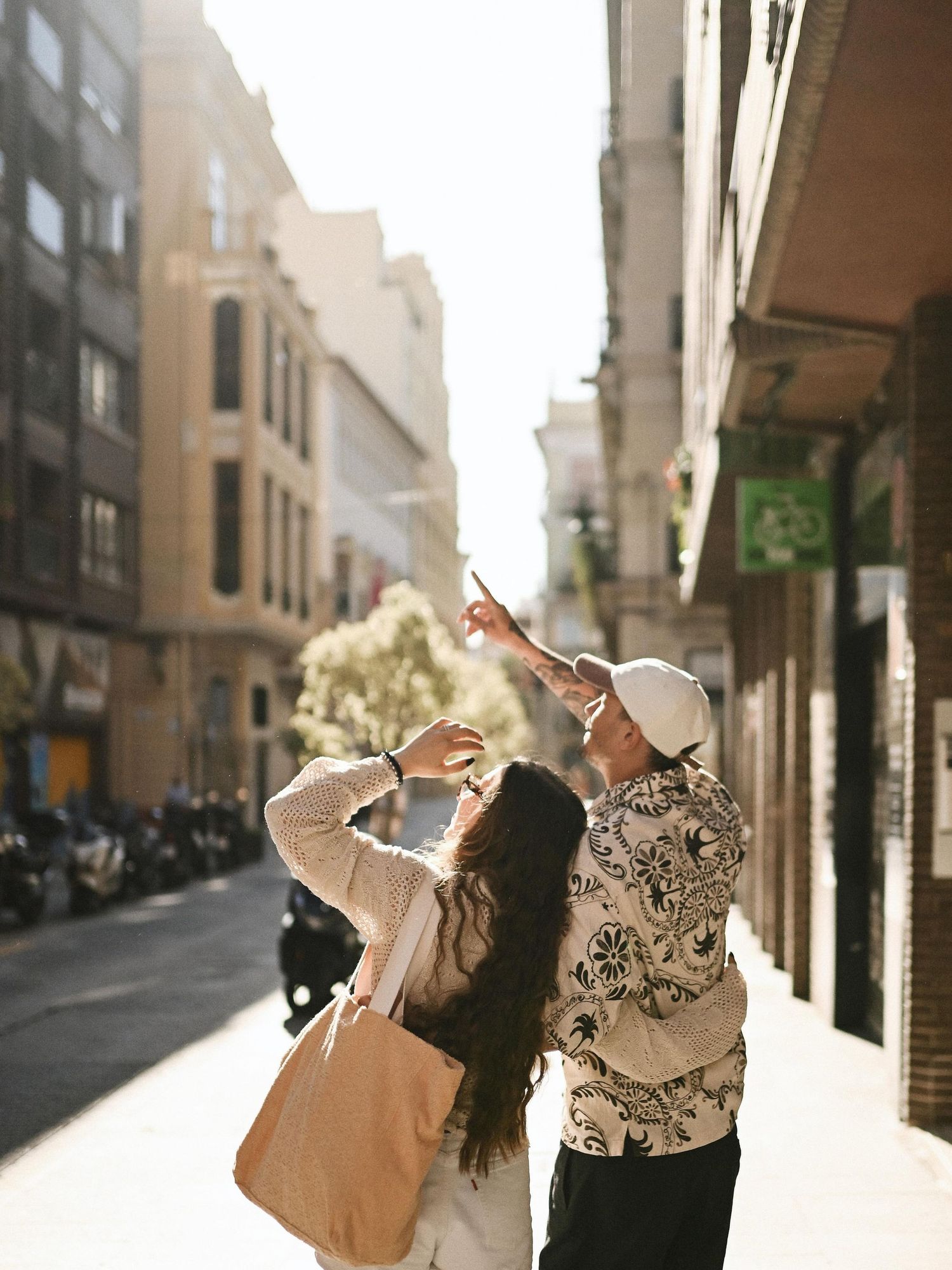 A couple seen from behind walking and pointing up at historic buildings along a sun-drenched street in Valencia center.
