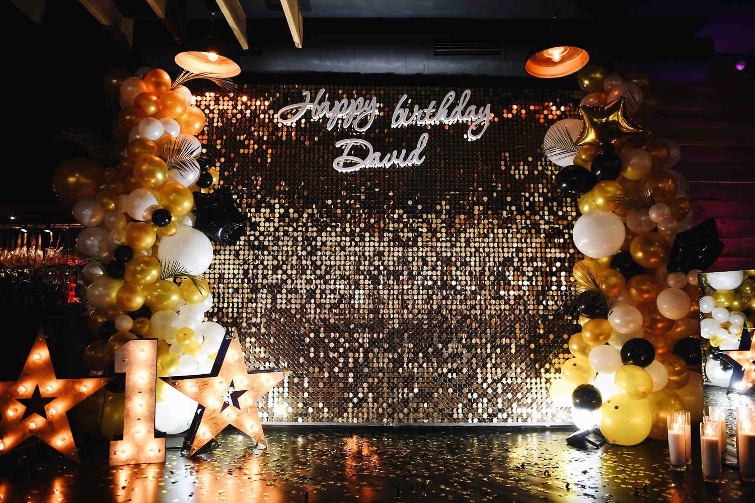 Empty luxury photo zone featuring a gold sequin wall, balloon arches, light-up marquee number 1, and glowing stars.