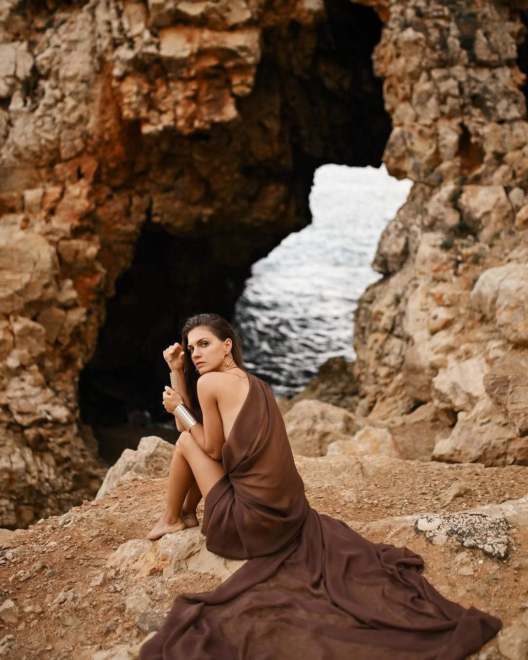 Woman in a draped dress sitting on the rocks during a professional portrait session at Cala Moraig.
