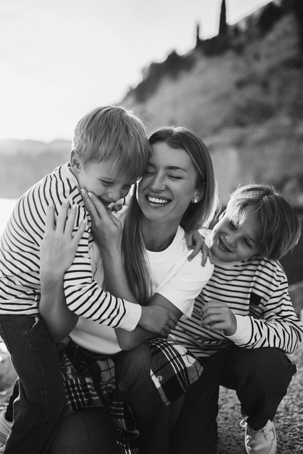 Candid black and white portrait of a laughing mother sharing a joyful, tight hug with her two sons on the beach in Calpe