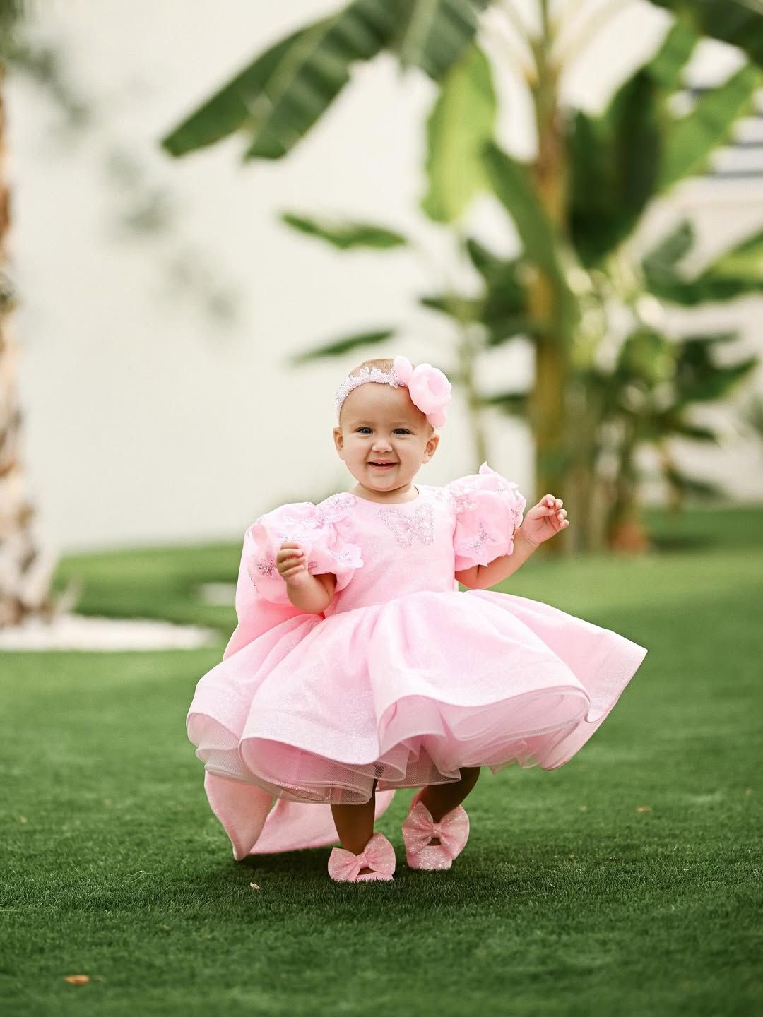 A laughing baby girl twirling in her pink dress on green grass with palm trees in the background during her birthday celebration.