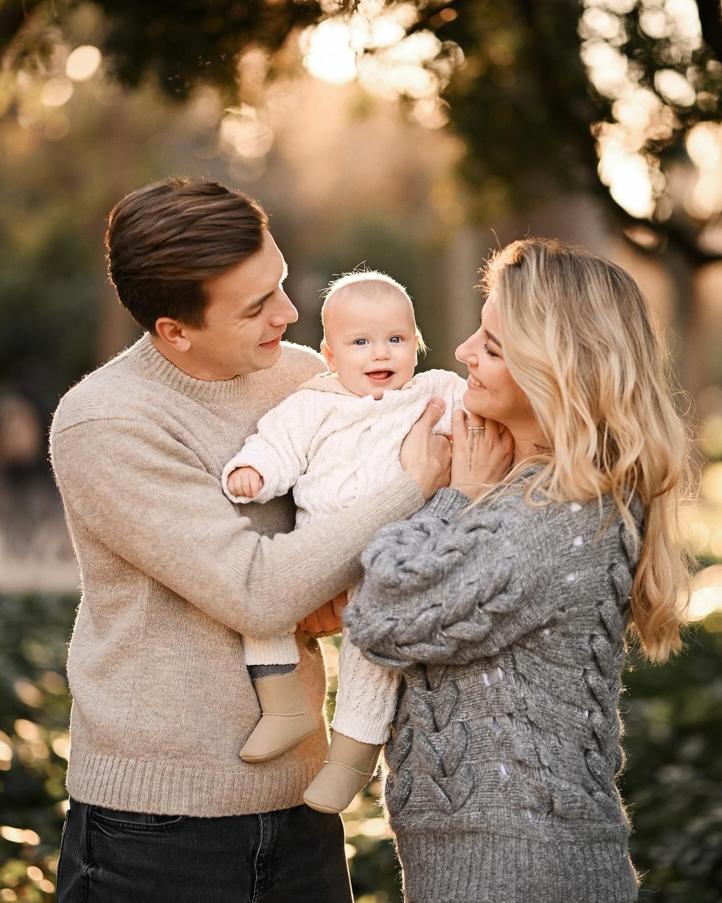 Happy mother and father in cozy sweaters holding and looking at their smiling baby during a family session in Turia Gardens.