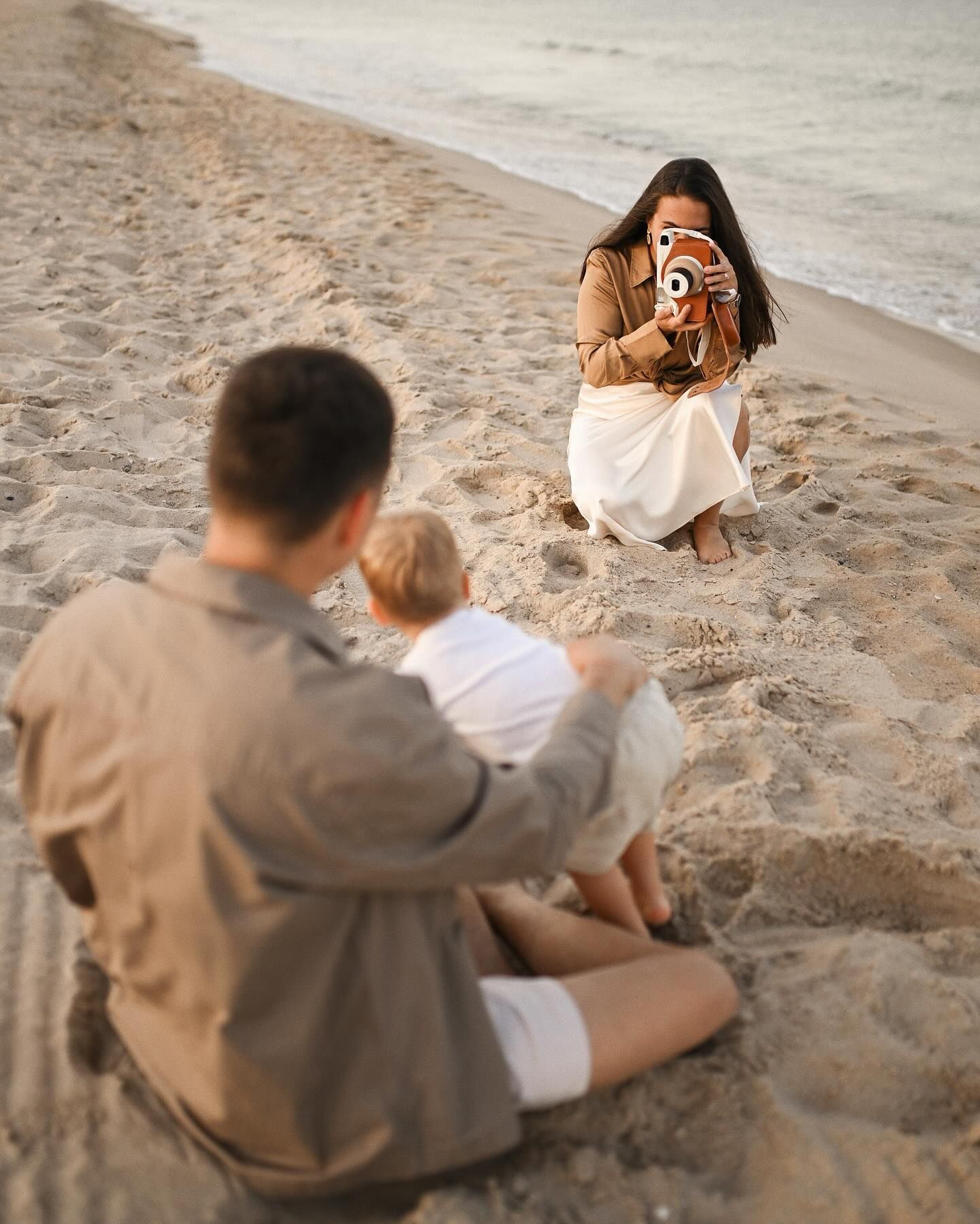 A mother sitting on the sand and using a vintage camera to photograph her husband and child during a candid beach session in Valencia.
