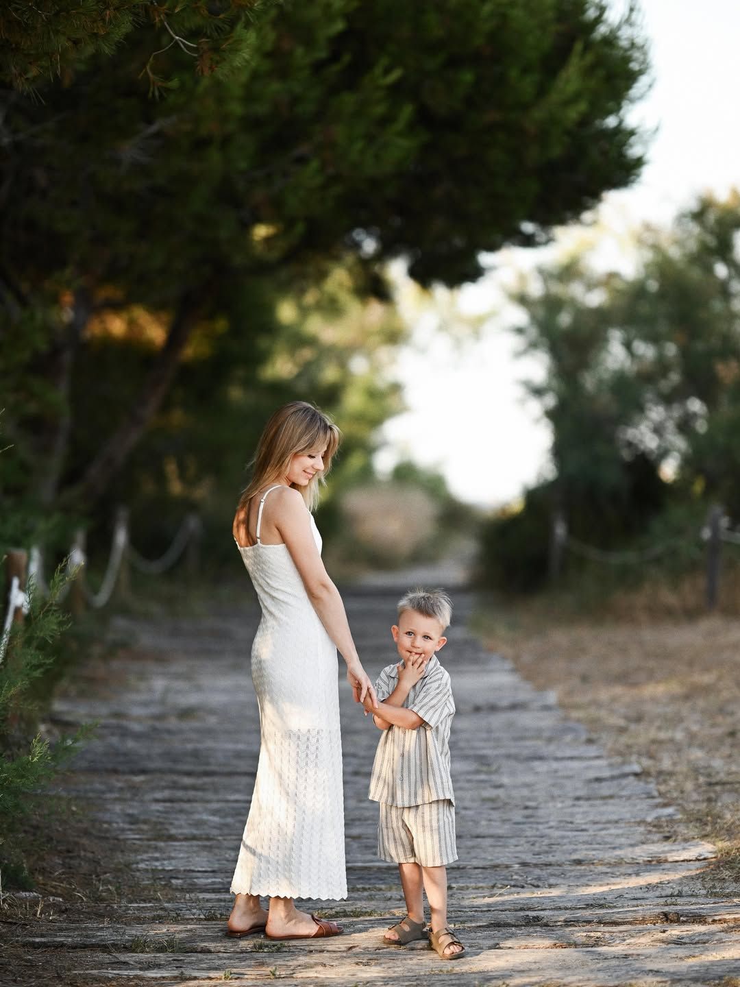 A mother holding her son's hand as they walk along a shaded wooden path among the pine trees of El Saler.