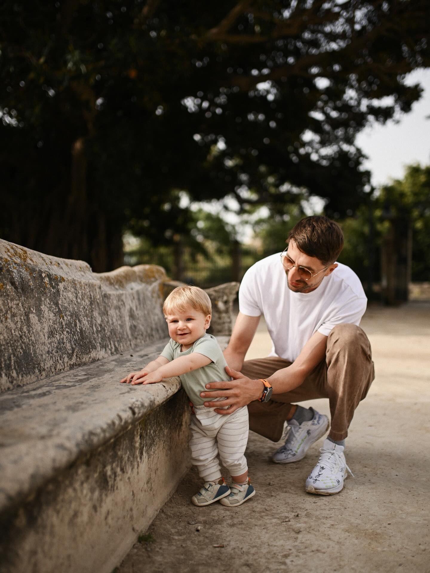 A cute baby boy balancing by an old stone bench with his father watching, captured during a sitter session in a Valencia park.