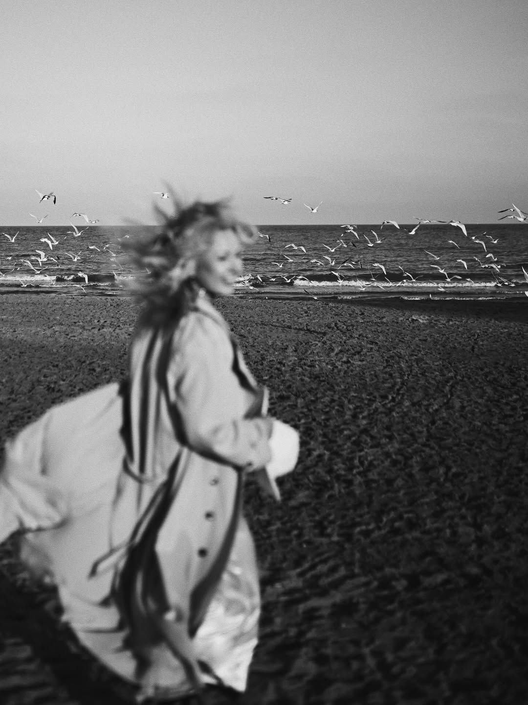 Black and white motion blur portrait of a blonde woman in a trench coat running on a beach with flying seagulls in Costa Blanca.
