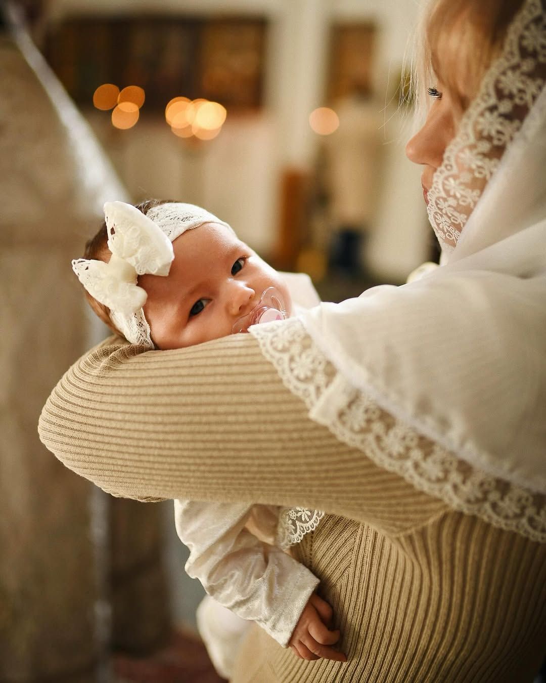Cute baby girl wearing a white lace bow held safely by her godmother during a quiet baptism service in Valencia