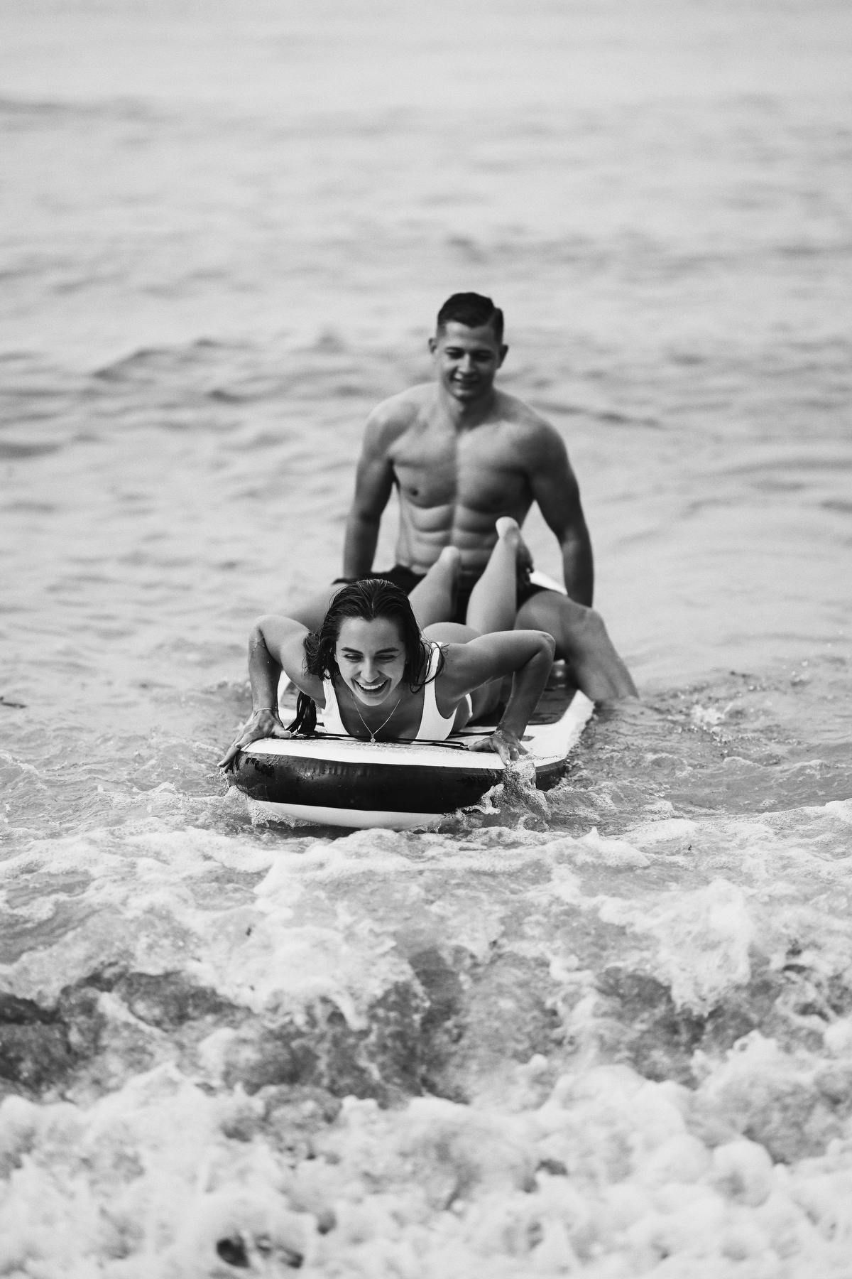 Playful black and white shot of a smiling woman on a paddleboard riding the waves with her partner in Moraira.