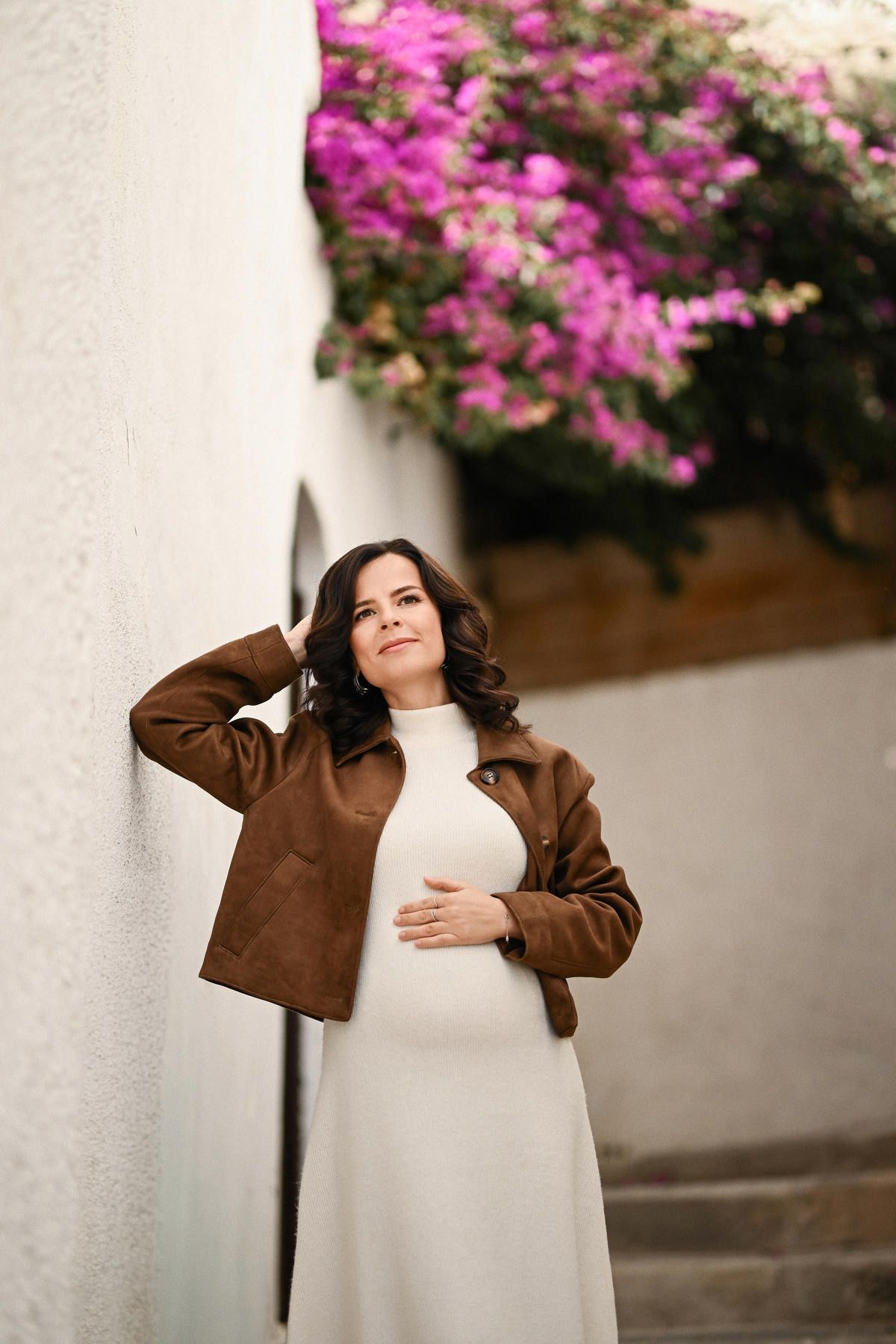 A beautiful pregnant woman in a white dress leaning against a traditional white wall with pink bougainvillea in Altea
