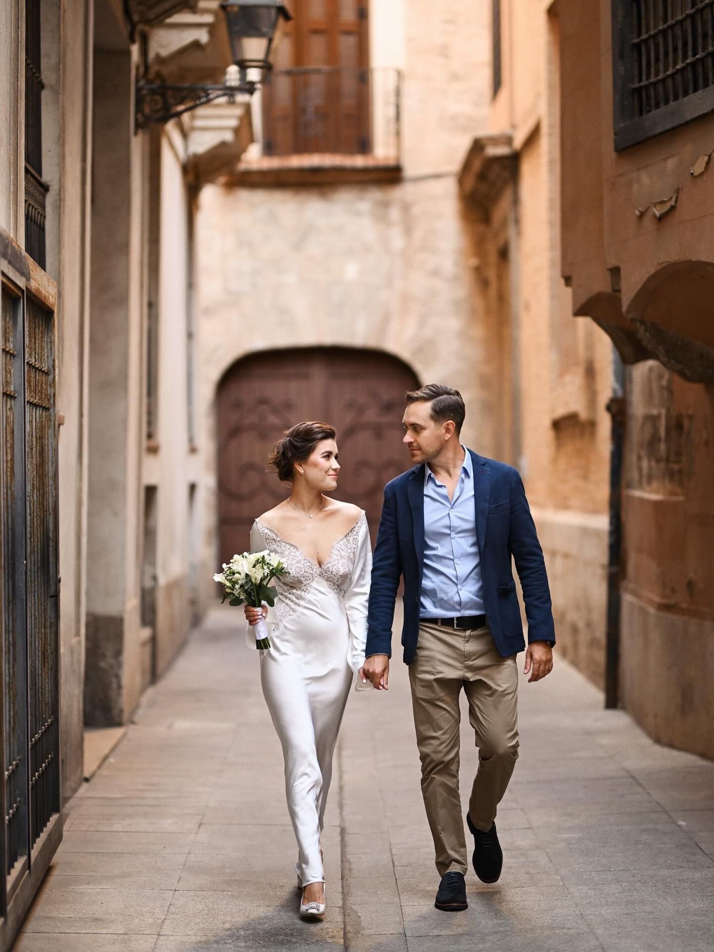 A bride and groom holding hands and sharing a look while walking down a narrow historic street in Valencia's El Carmen district.