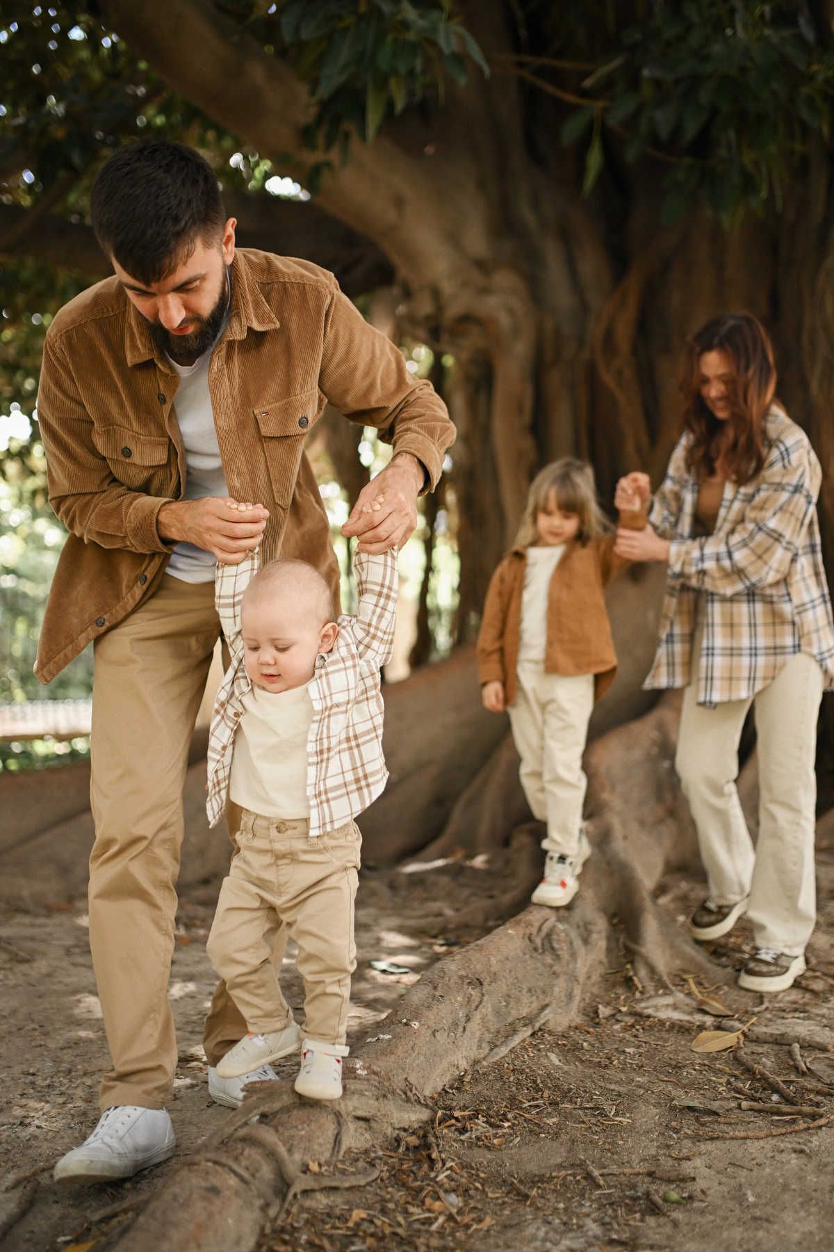 Father helping his baby boy balance on large tree roots while mother and sister walk behind in Jardines del Real.