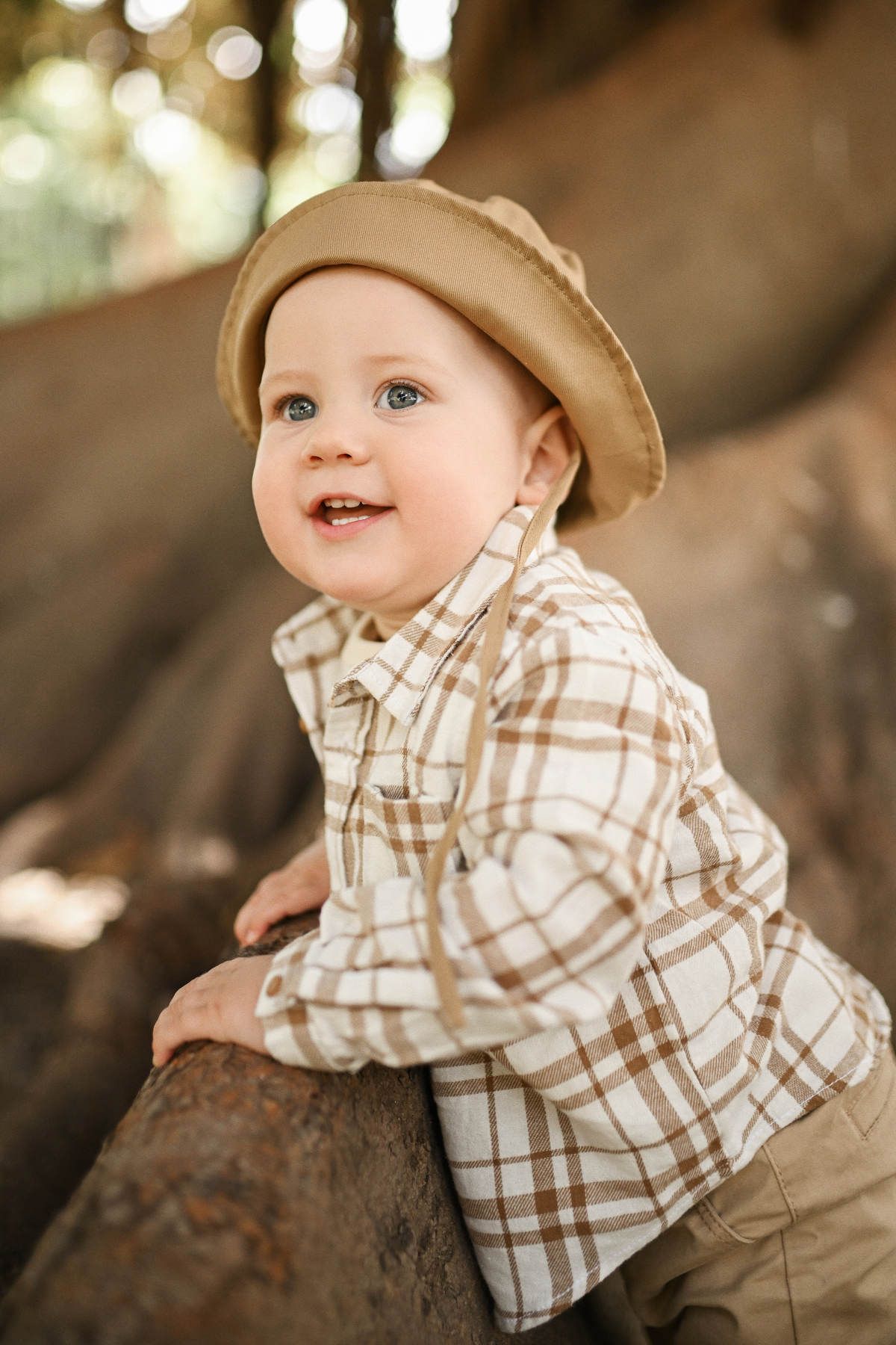 A cheerful baby boy in a bucket hat leans against a massive ancient tree root exploring nature in Jardines del Real.