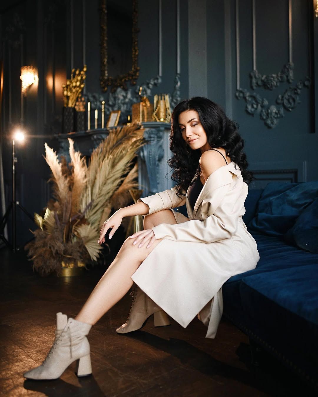 Cinematic boudoir photoshoot in a Valencia studio. A woman in a beige coat sits elegantly on a blue velvet sofa with moody lighting.