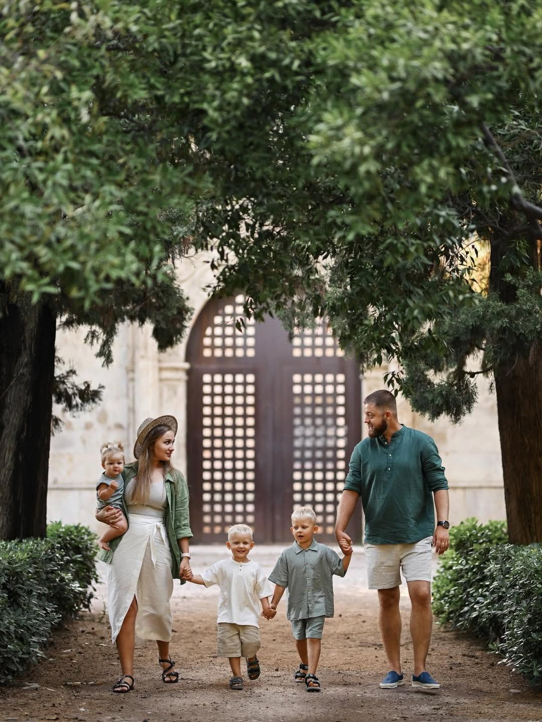 A family walking hand-in-hand along a shaded path toward an old wooden gate in Turia Gardens, Valencia.