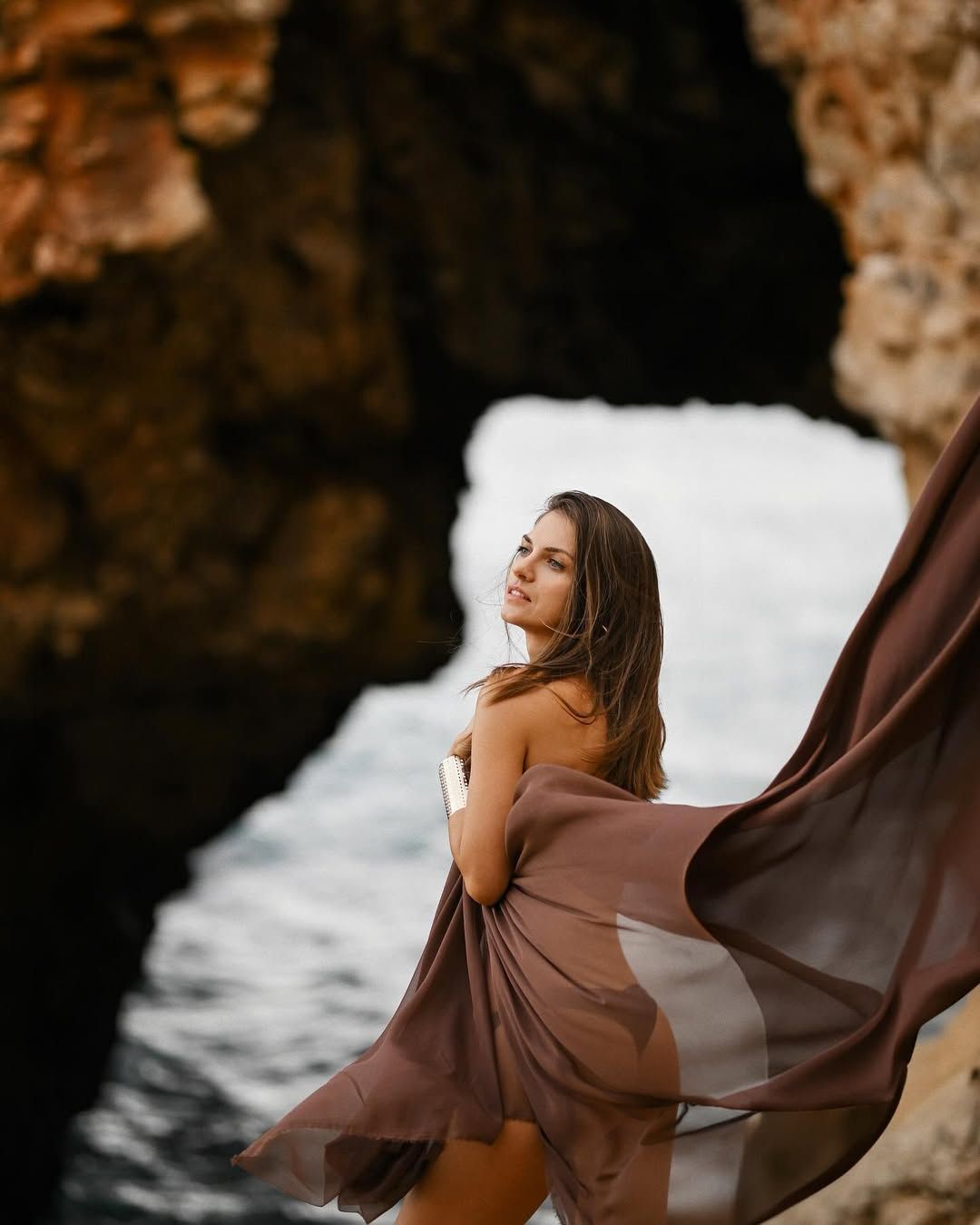 Woman in a flowing brown dress posing against limestone cliffs during a portrait photoshoot at Cala Moraig.