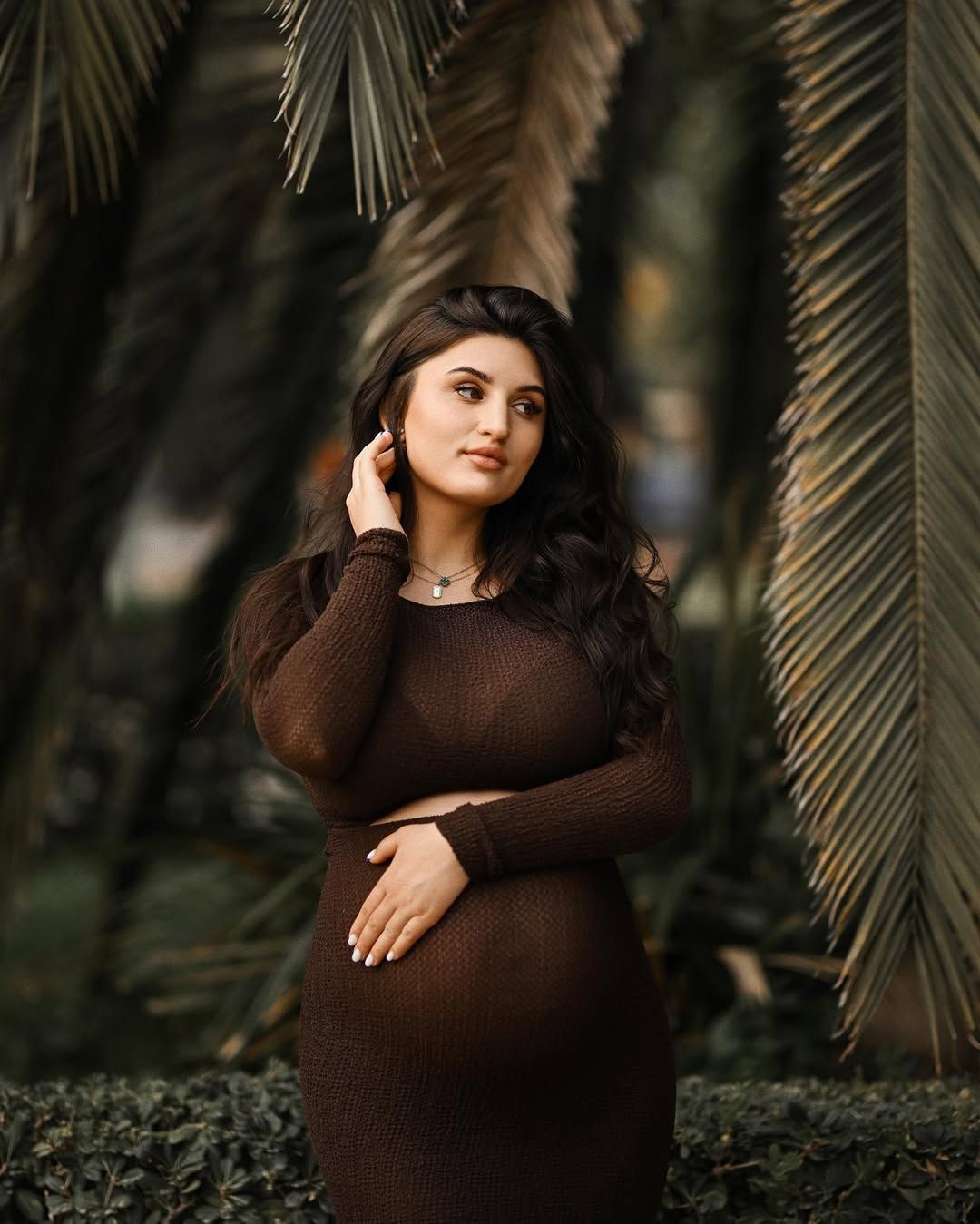 Pregnant woman gently holding her belly next to large green palm fronds during an elegant maternity photoshoot in Viveros Park.