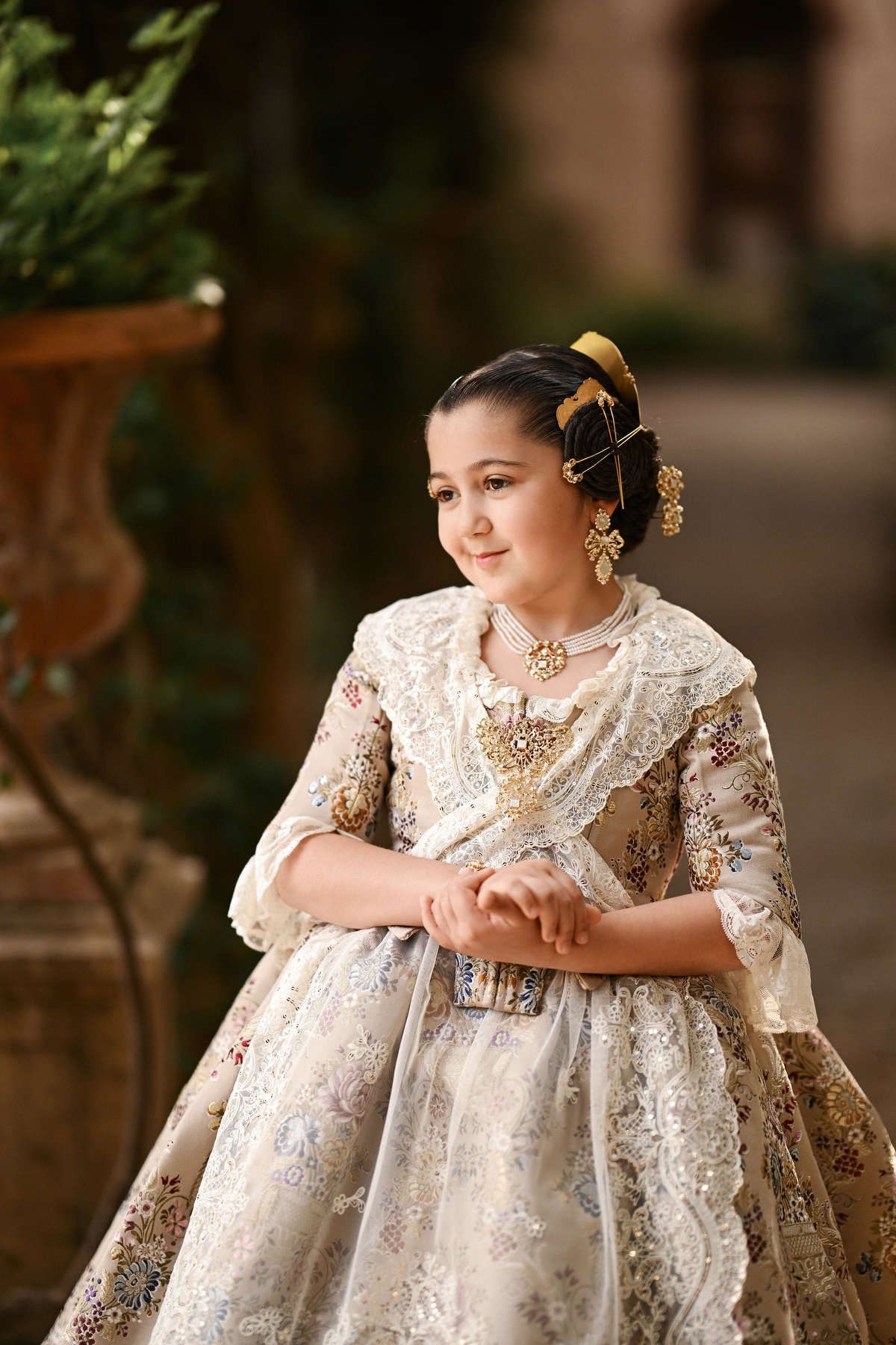 A portrait of a young girl in a bespoke silk Fallera dress, posing elegantly with her hands clasped in the tranquil Jardines de Monforte.