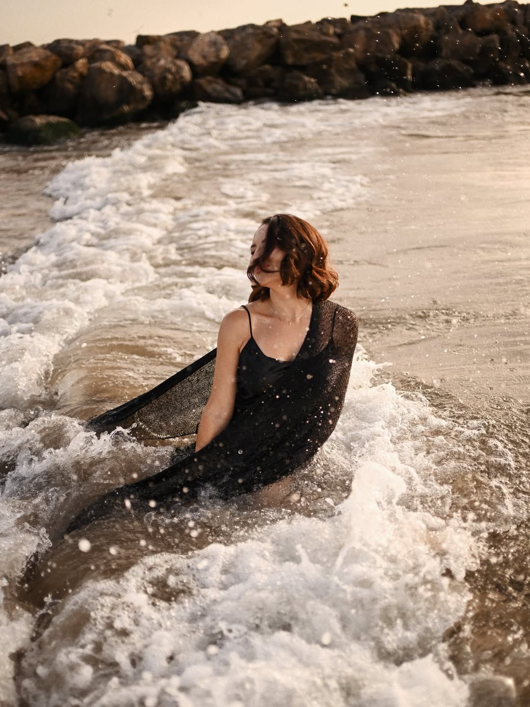 Editorial women's portrait in Valencia, sitting in crashing ocean waves in a black dress with water splashing around her.