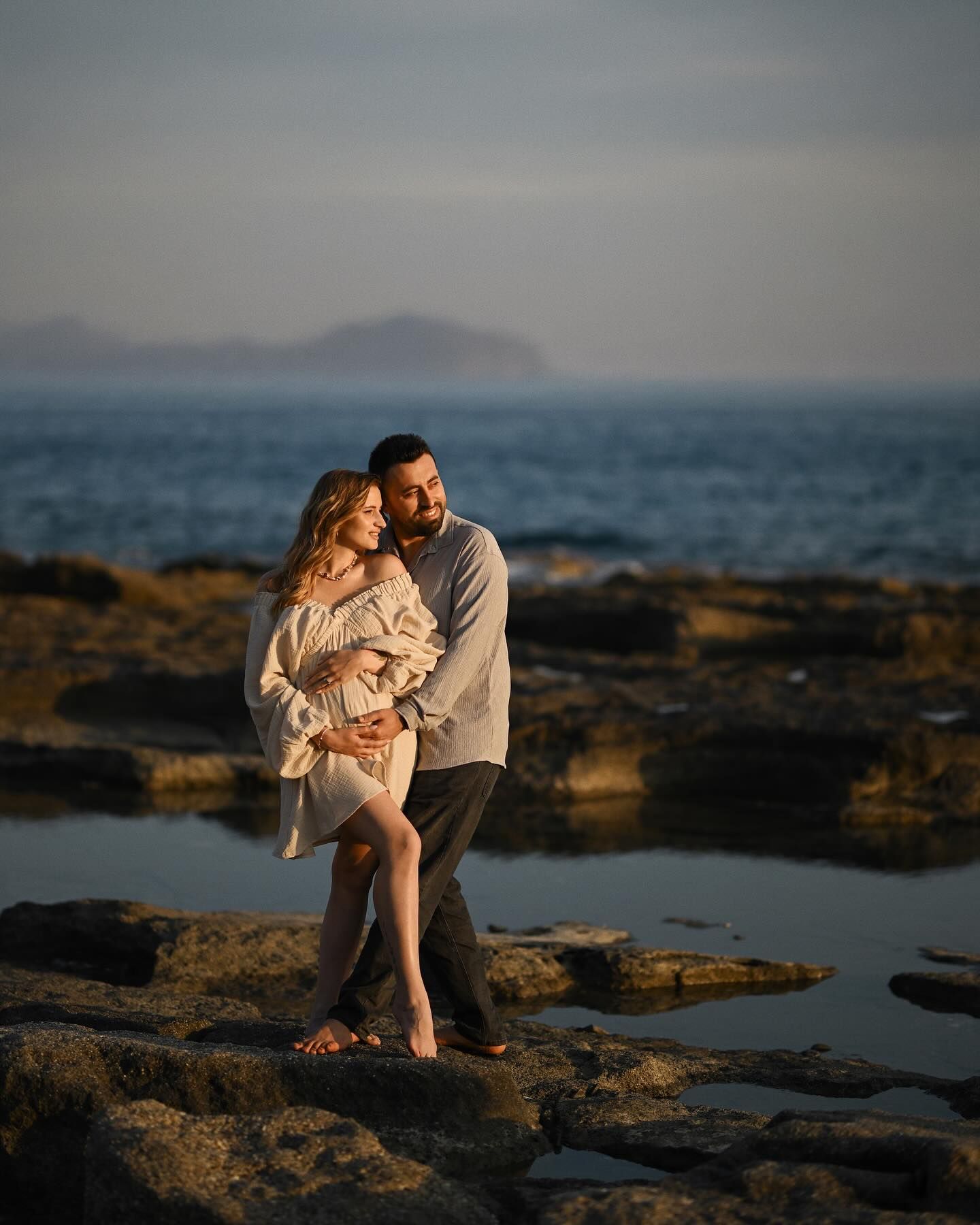 Full-length portrait of a husband embracing his pregnant wife on a rocky shore during a golden hour session in Costa Blanca.