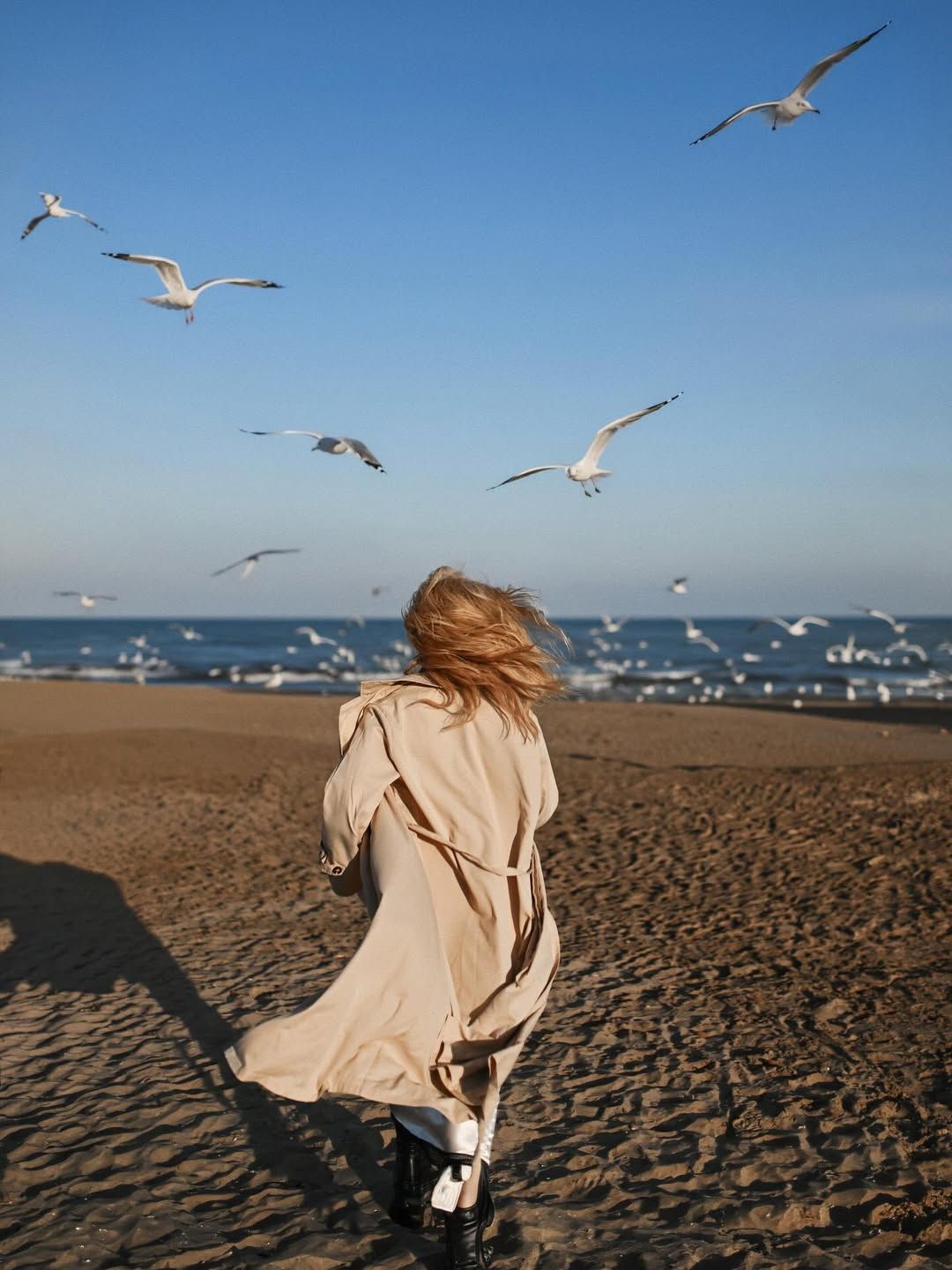 Full-length back view of a blonde woman in a trench coat walking along the shoreline under flying seagulls on a Costa Blanca beach.