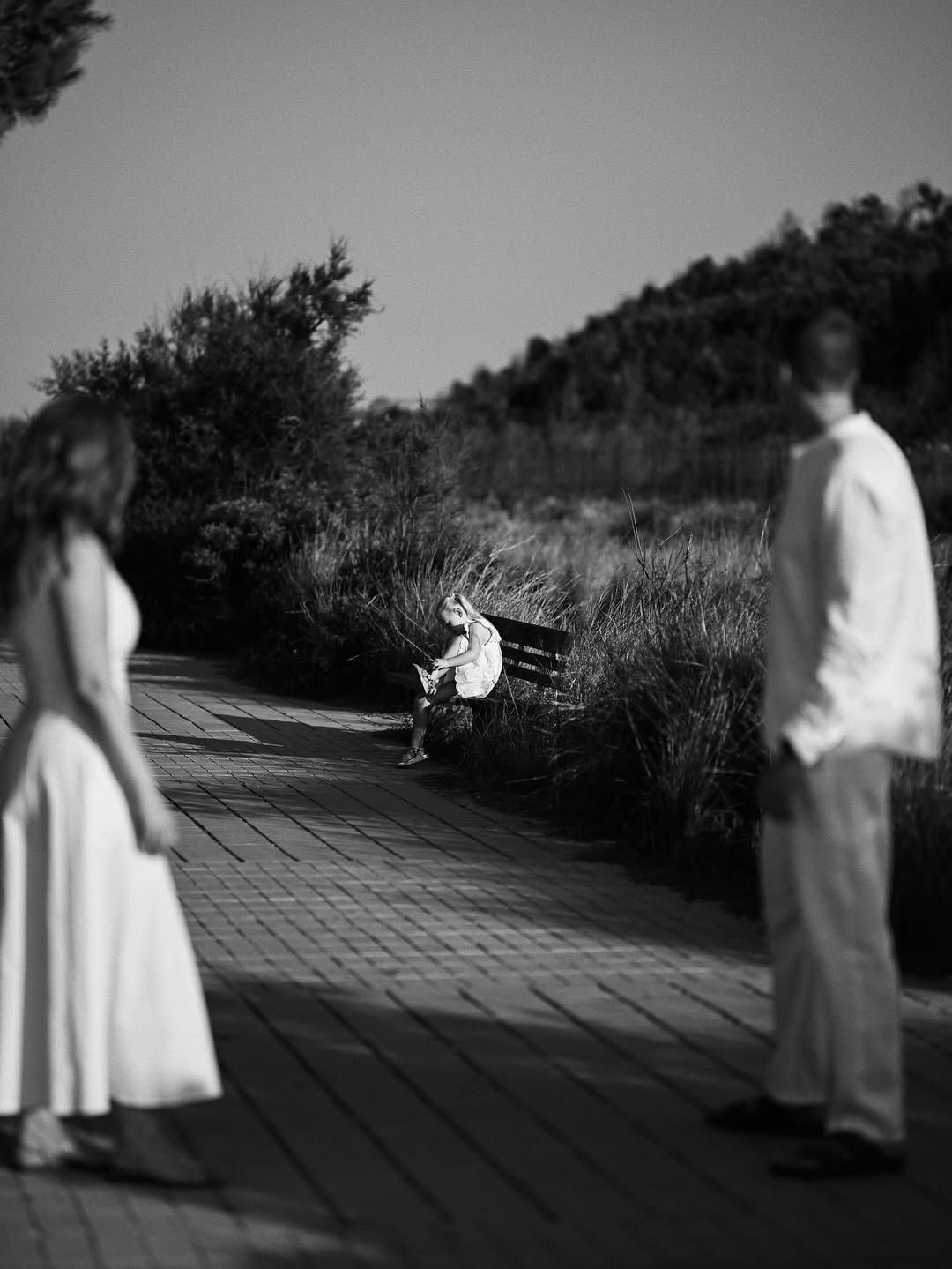 A little girl in a white dress sits alone on a wooden park bench in El Saler while her parents watch nearby.