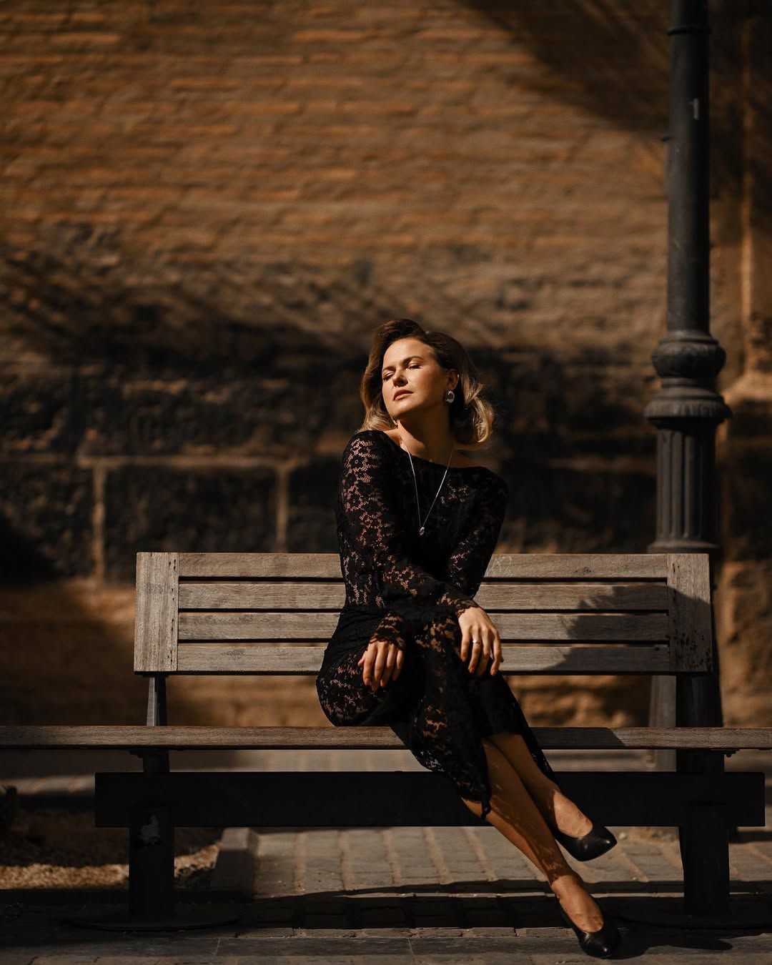 A beautifully styled woman in a black dress sits on a wooden park bench, lit by warm sunlight during an outdoor session.