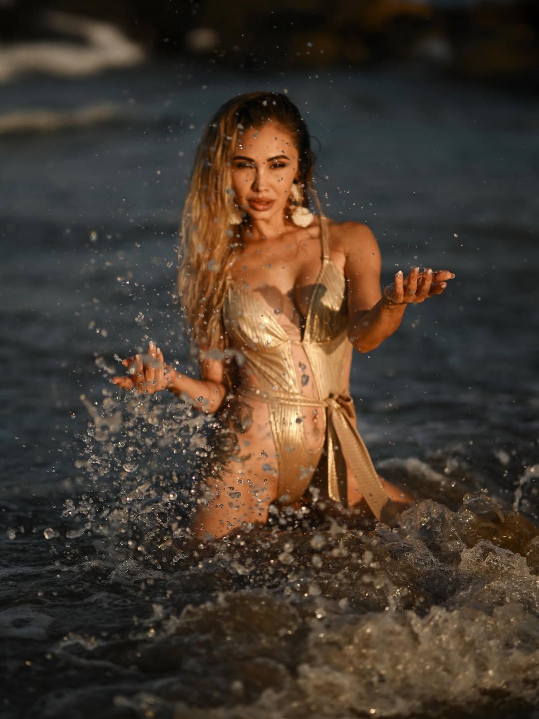 A woman in a gold swimsuit stands in the sea, creating a cinematic splash during an outdoor portrait session on a Valencia beach.