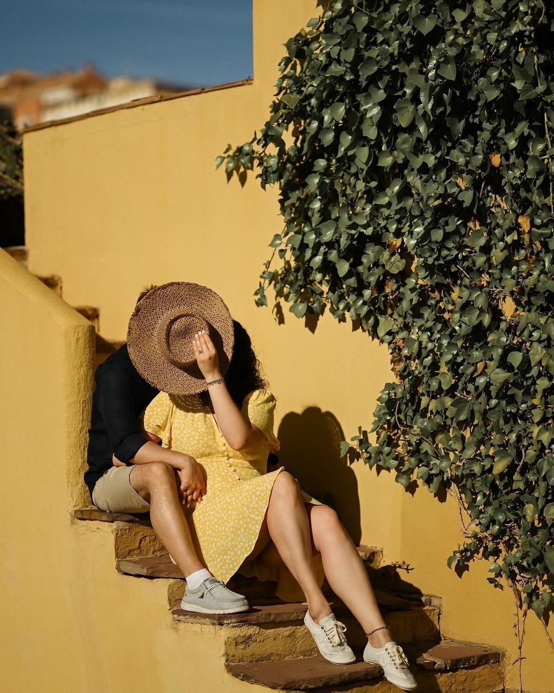 A romantic couple sits on the villa stairs, hiding their faces behind a hat against a mustard wall in Port Saplaya, Valencia.