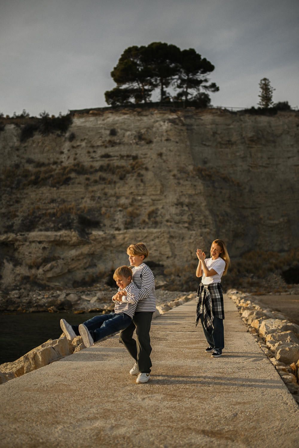 A mother smiling and clapping as her two boys play and spin on a stone pier with dramatic cliffs in the background in Calpe.