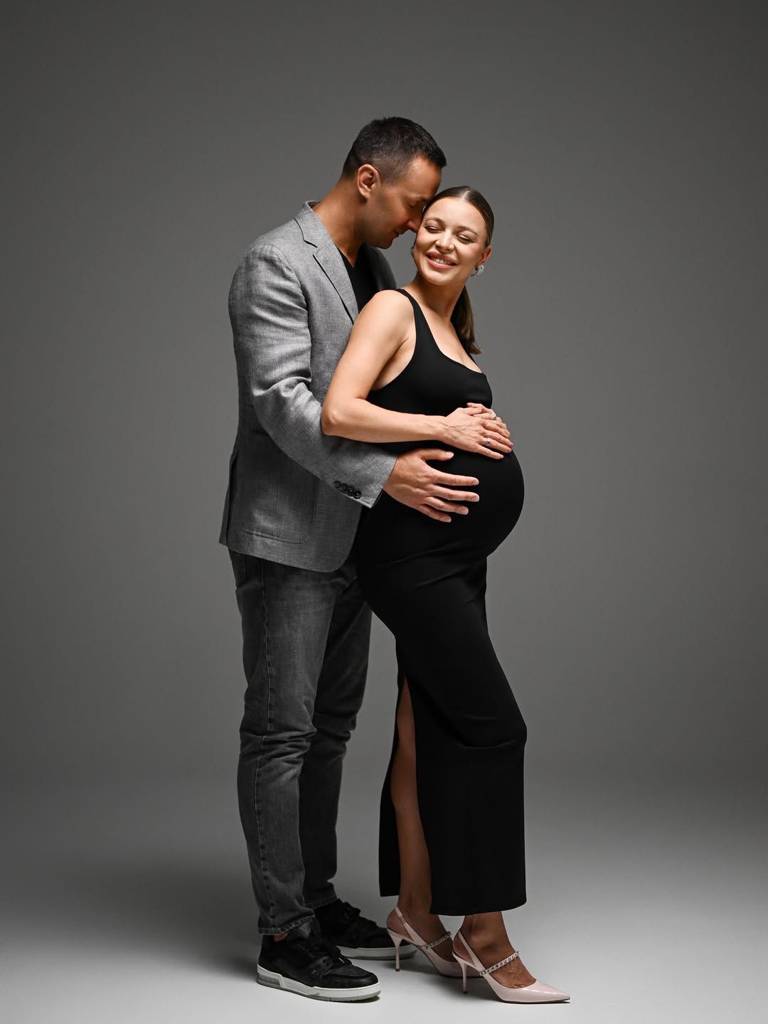 An elegant studio portrait of a couple expecting a baby, showcasing a tender and stylish moment against a grey backdrop