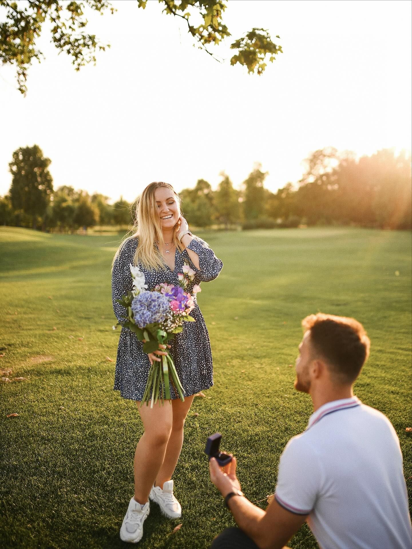 A man kneeling on the grass and proposing to a smiling woman holding a bouquet of flowers during a sunset outdoor session.