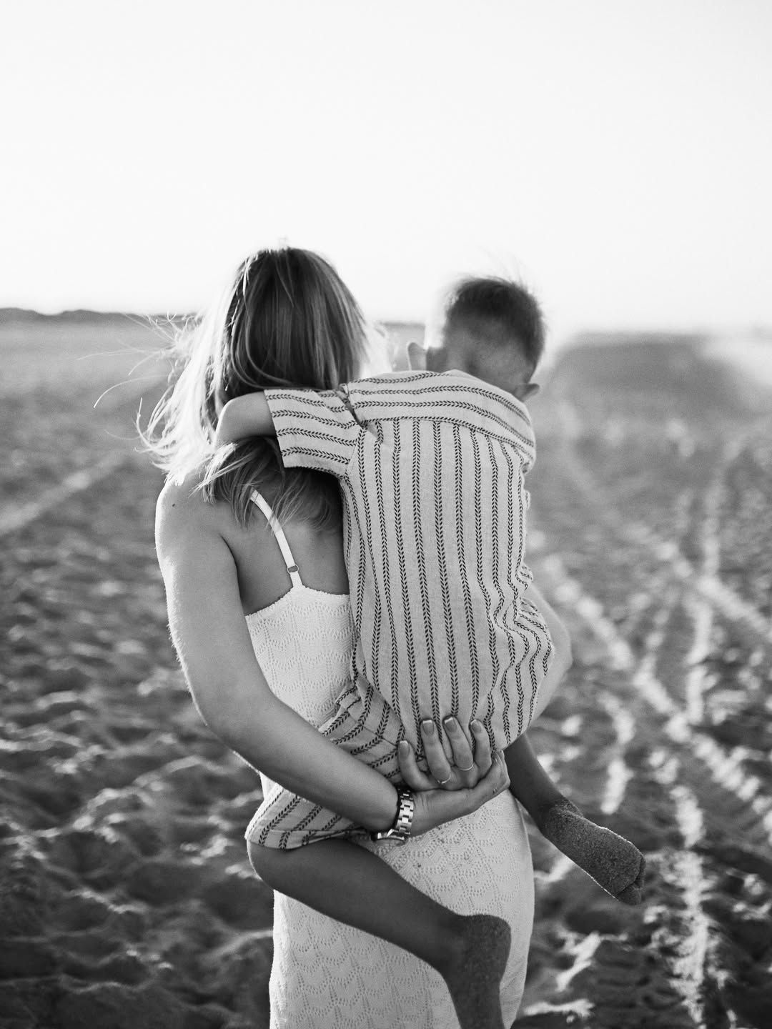 Black and white view from behind of a mother carrying her little boy across the textured sand of El Saler.