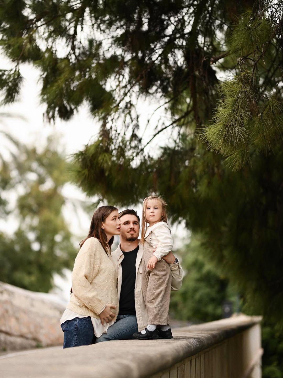 Expecting couple with their daughter during a maternity session in Turia Park, Valencia, surrounded by soft greenery and gentle shade