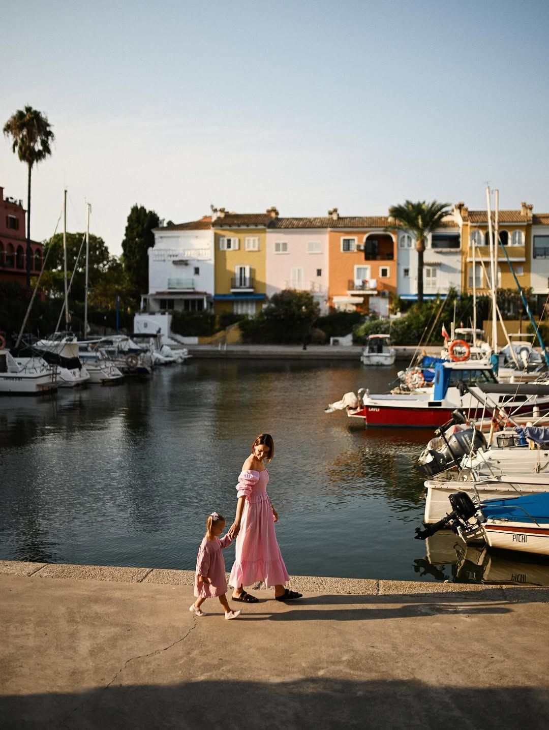 Wide shot of a mother and daughter walking along the waterfront with a scenic view of boats and colorful houses in Port Saplaya