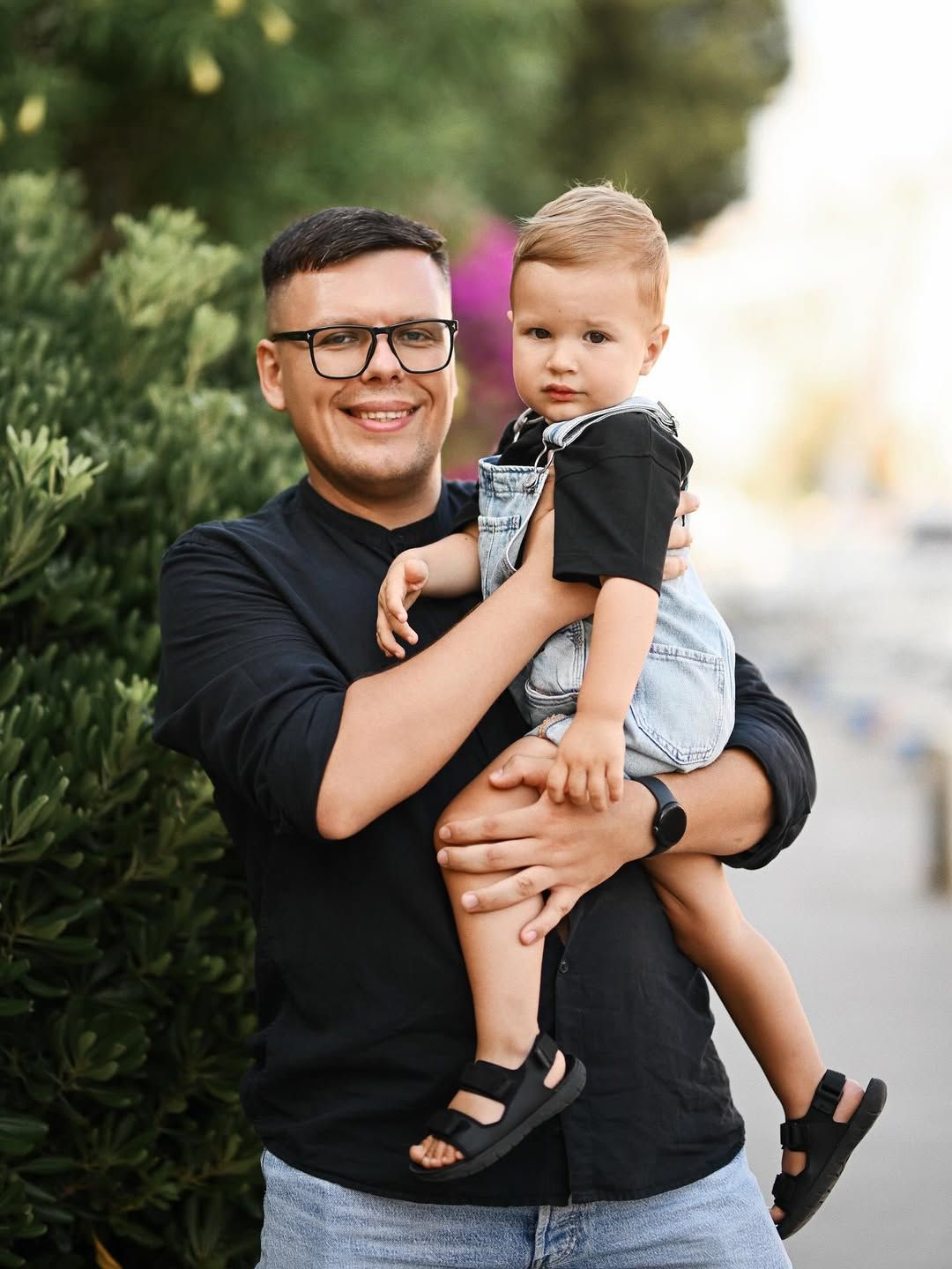 A father holds his young son on his arms, both looking directly into the camera during a family photoshoot in Port Saplaya