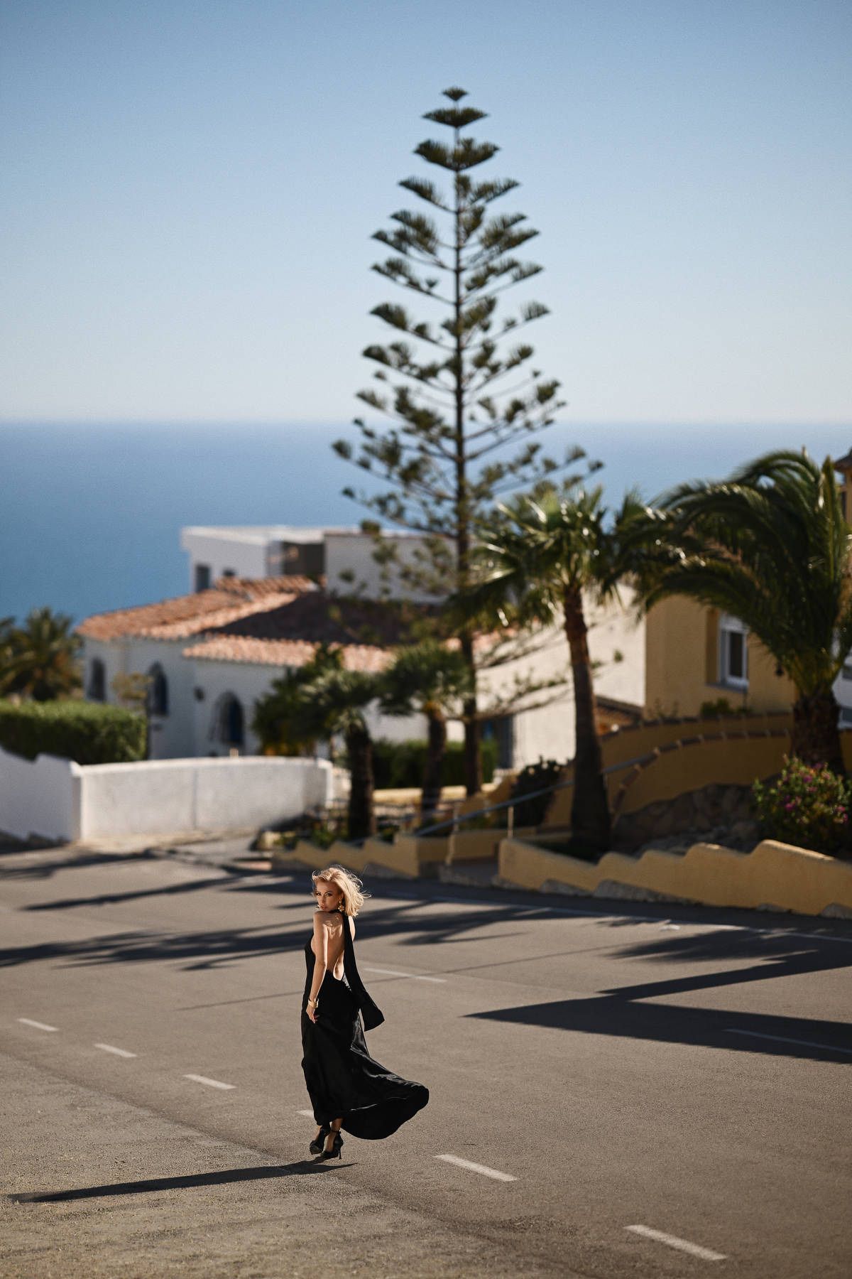 A woman walking along a quiet street lined with lush Mediterranean pines and white villas during a lifestyle session in Javea.