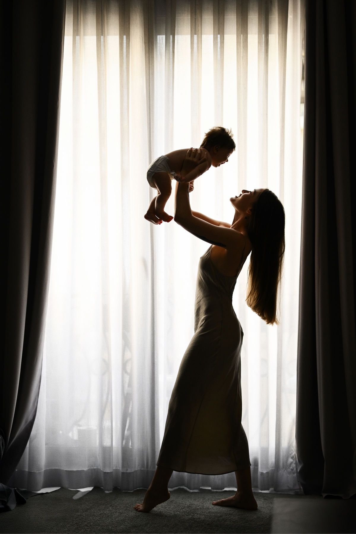 A beautiful silhouette of a mother lifting her baby high against a bright window, highlighting a joyful motherhood connection.