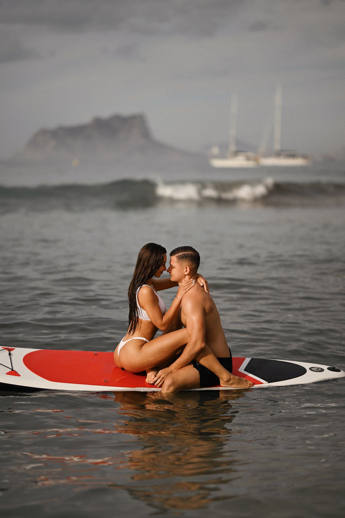 A couple sitting together on a paddleboard in the Mediterranean Sea with the Moraira coastline in the background