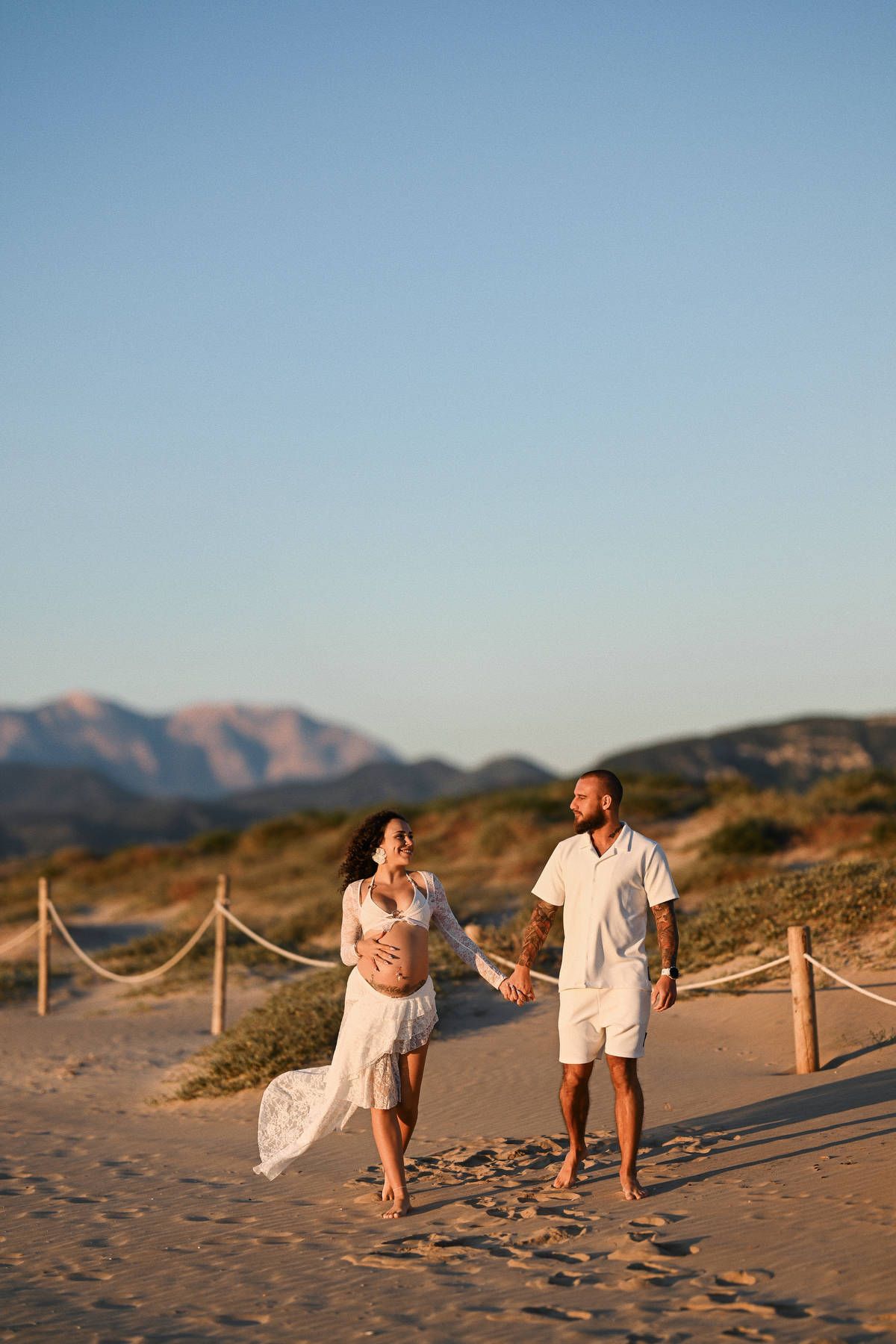 A happy pregnant woman and her partner holding hands and walking along the beach with mountains in the background.