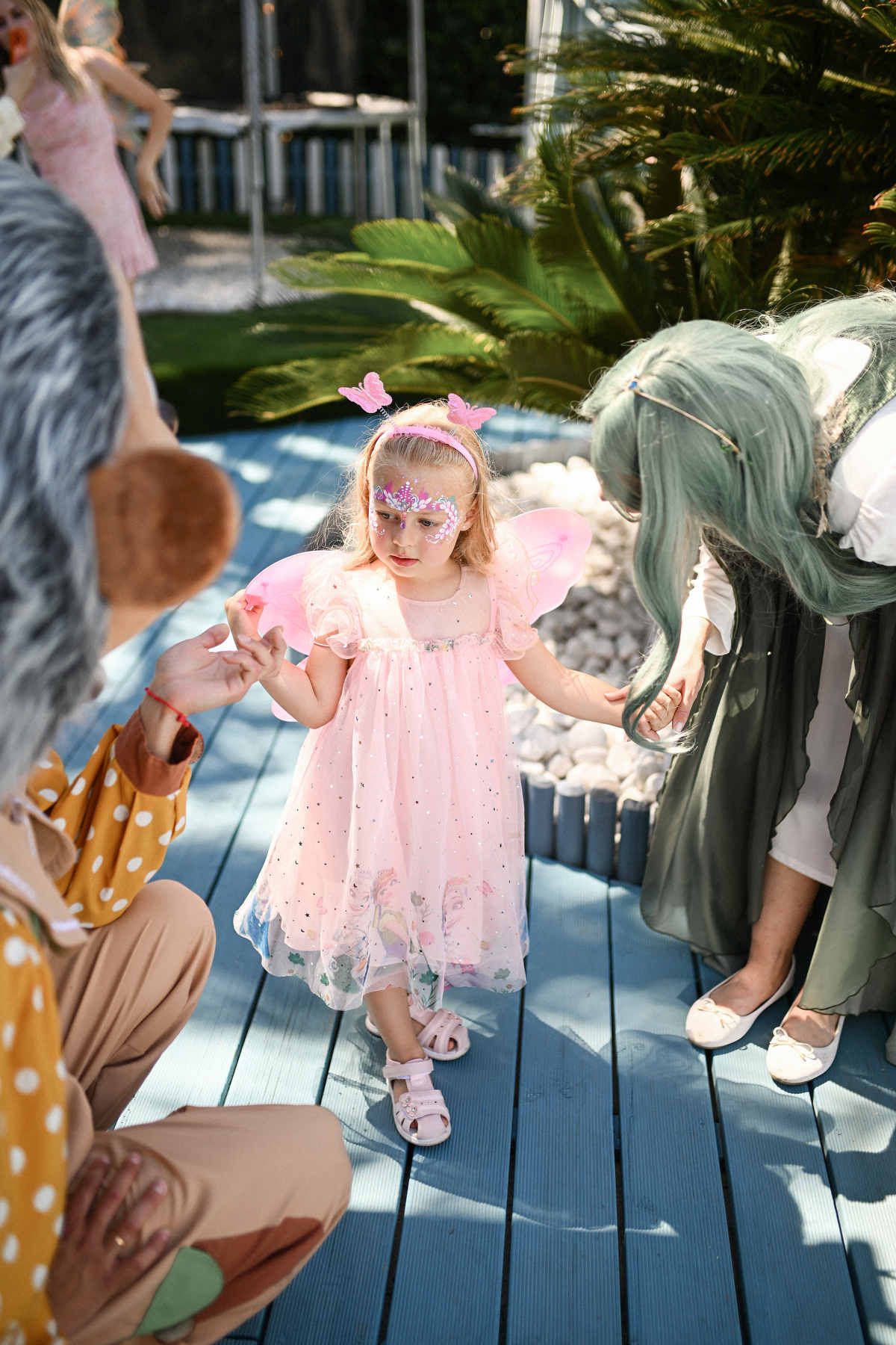 A little girl wearing pink fairy wings talking to interactive costume characters during a children's birthday entertainment show.