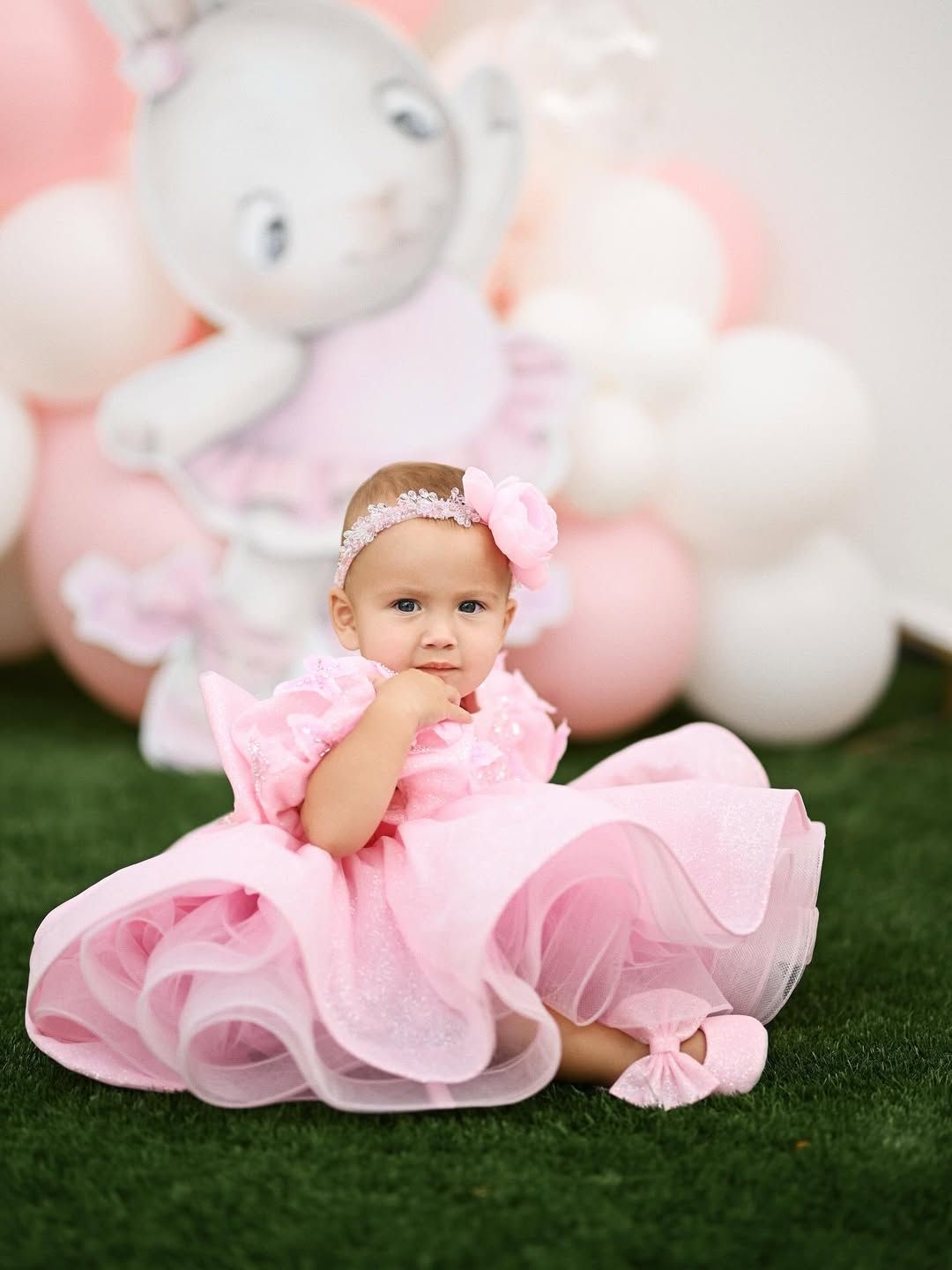 Toddler girl in a pink dress and floral headband sitting on green grass in front of birthday party balloons.