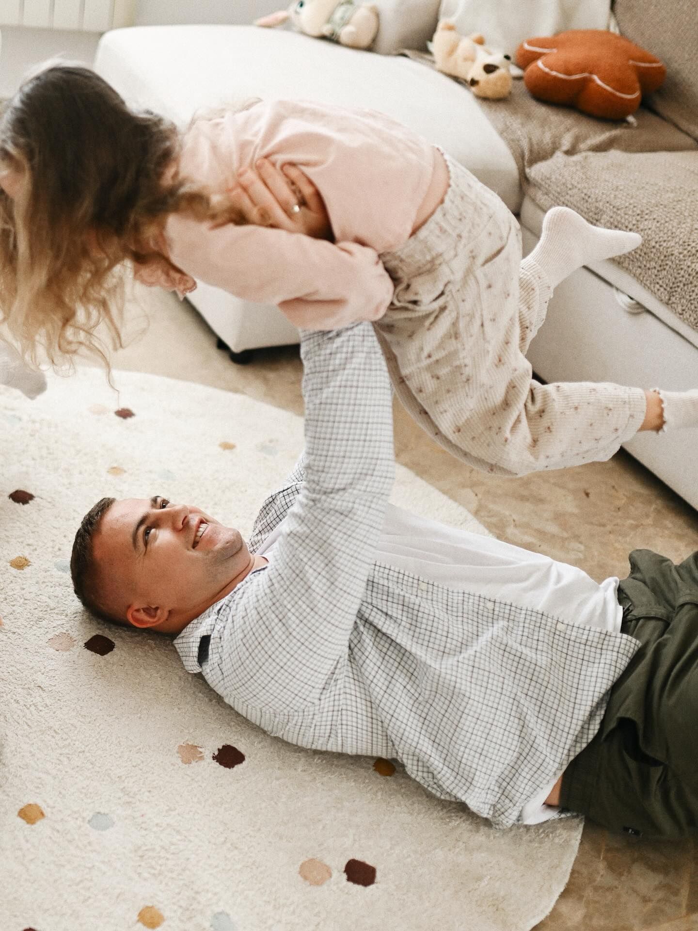 A joyful father lying on a rug and playfully lifting his young daughter high into the air during a candid family home photo session.