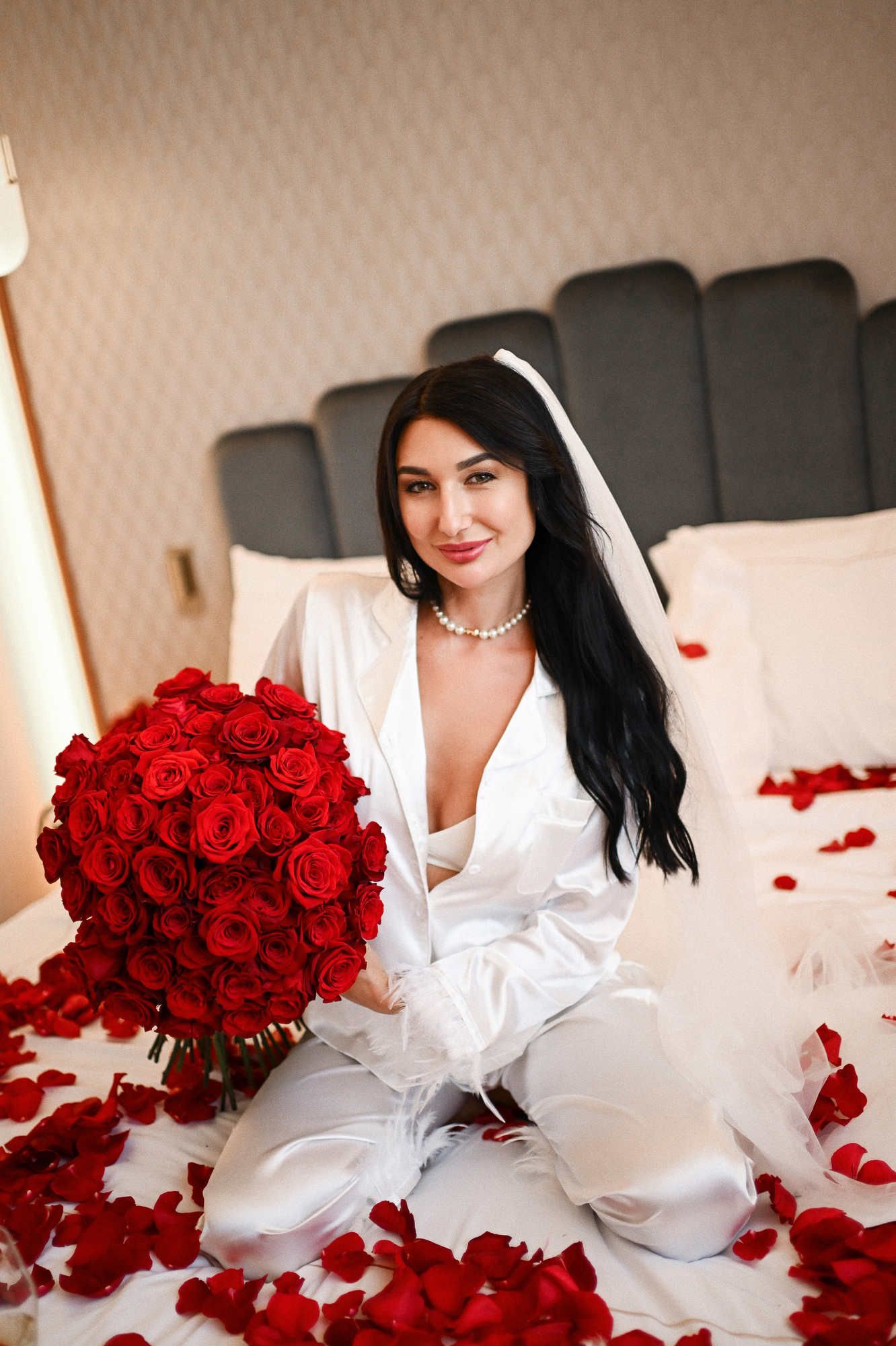 Bride's first look during a hen party photoshoot in Valencia, wearing white silk pyjamas and holding red roses on a bed.