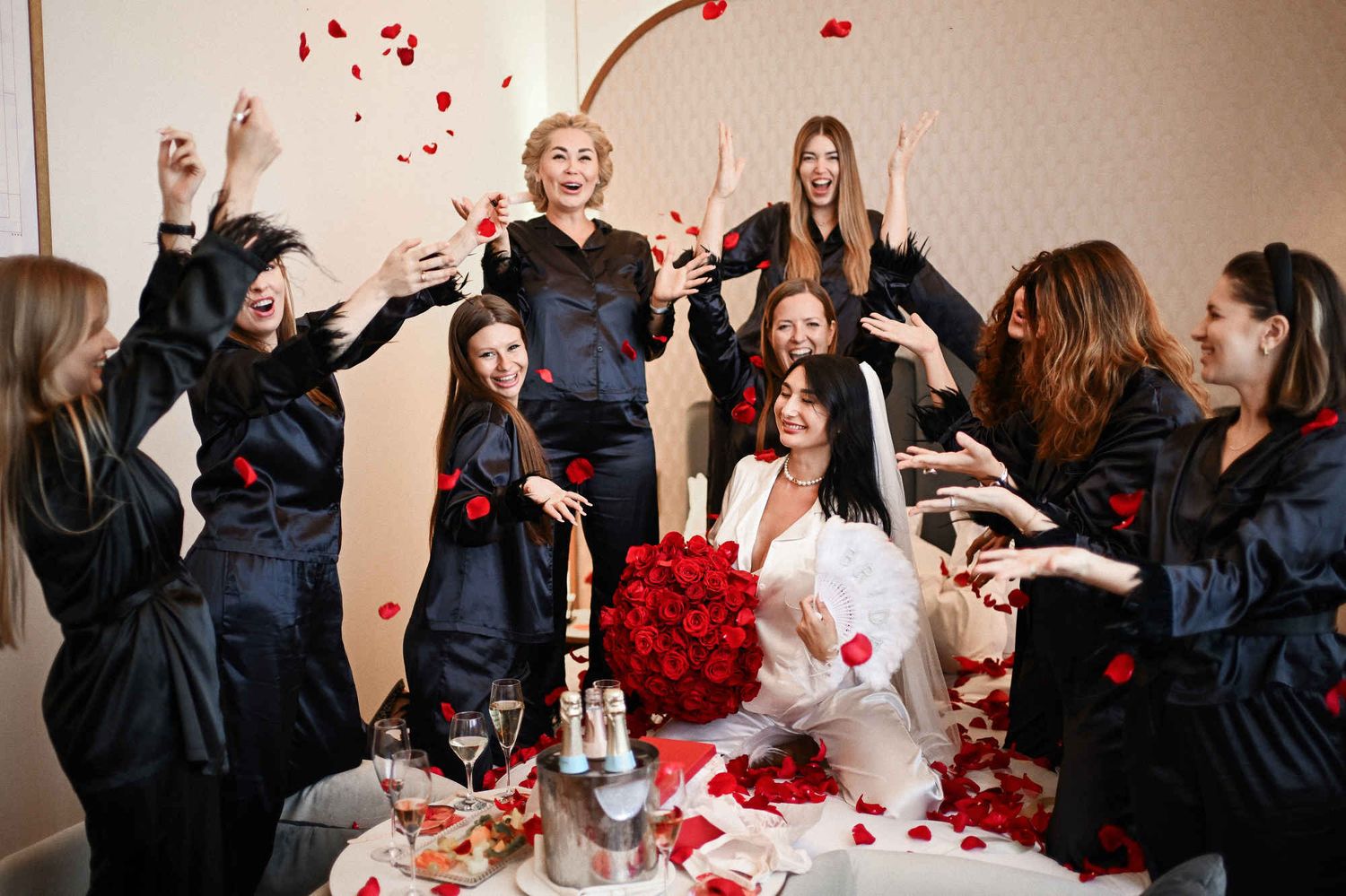 Bridesmaids in black pajamas throwing red rose petals over the bride holding a large bouquet in a Valencia hotel room.