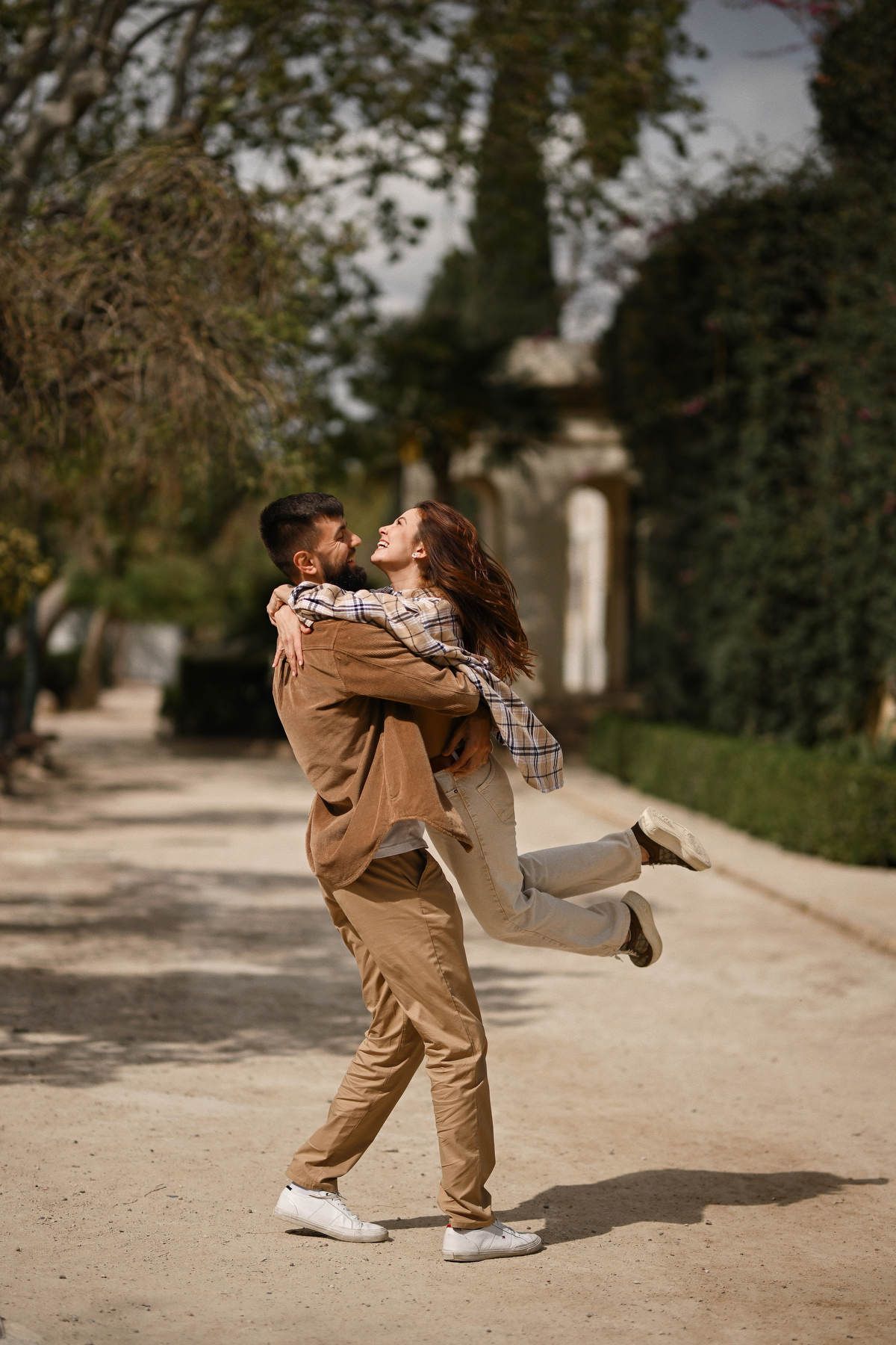A father playfully lifts his laughing wife in the air on a wide dirt pathway during a family shoot in Jardines del Real.