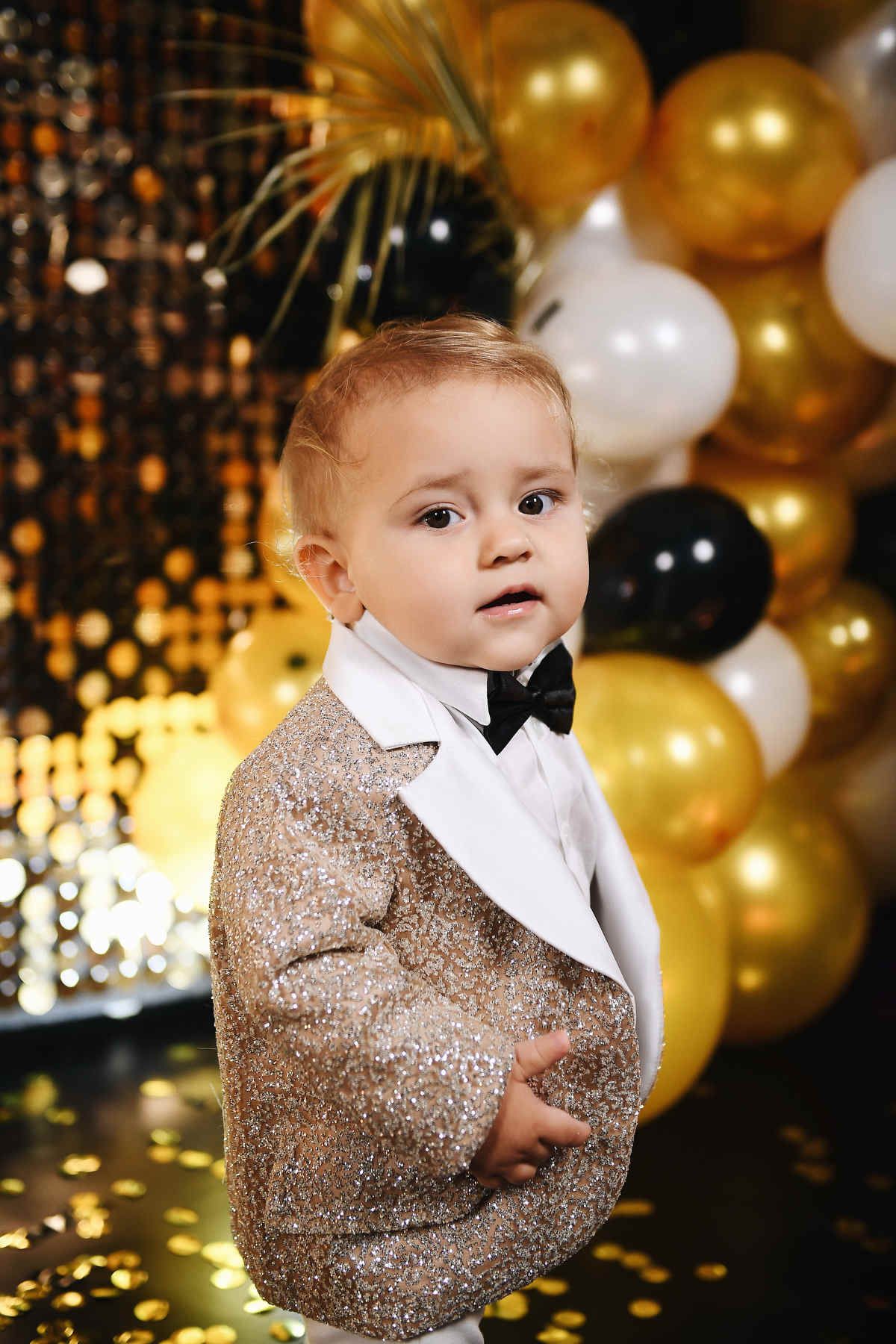 Close-up portrait of a one-year-old boy wearing a sparkling gold blazer and black bow tie at his birthday celebration.