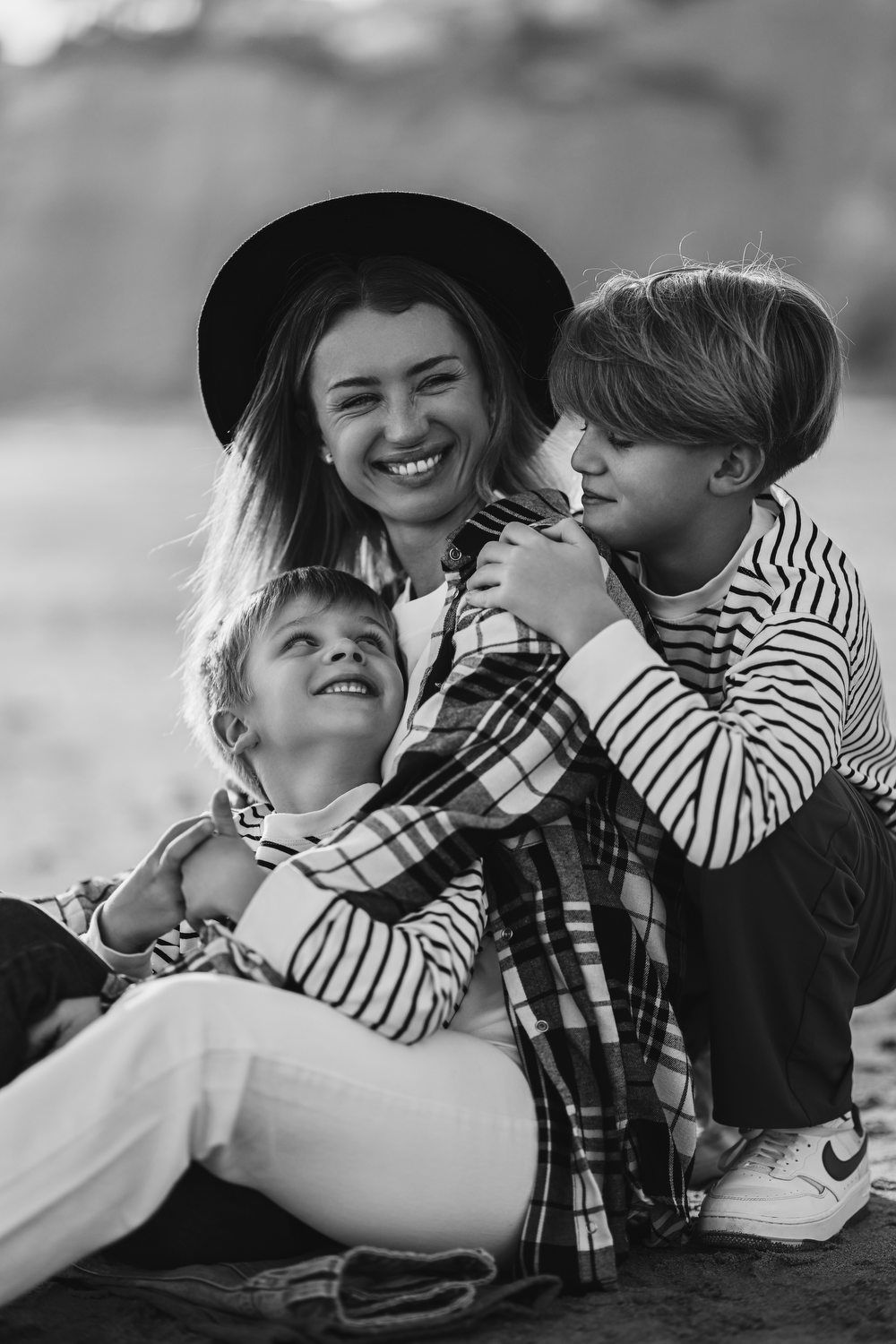 A joyful black and white shot of a mother laughing as her two boys hug her affectionately while sitting on the sand in Calpe